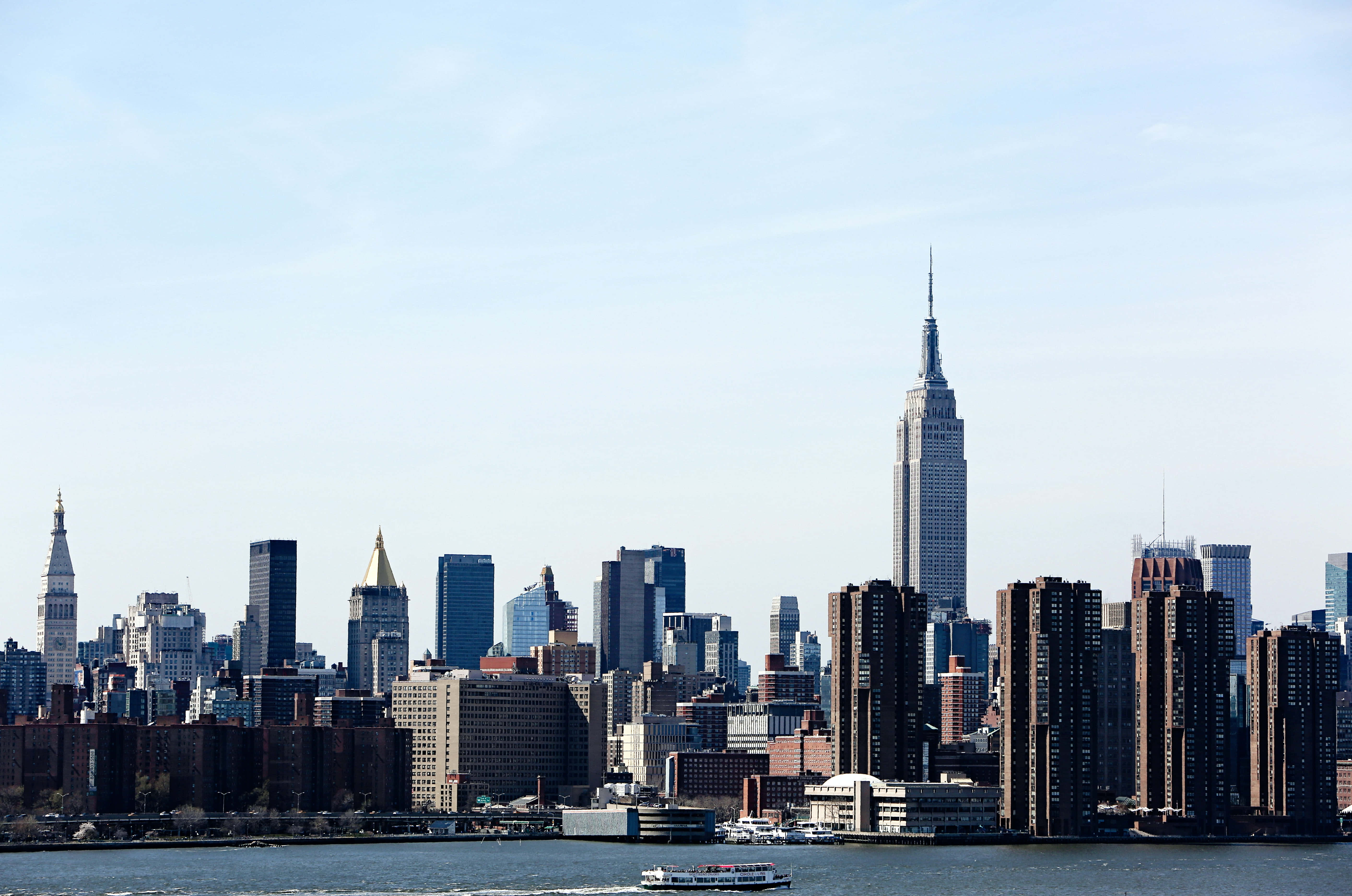 Nyc 桌布 壁紙 — 紐約市天際線的明亮日間景觀，帝國大廈聳立於現代和歷史摩天大樓之上，旁邊有一條河流和渡輪。