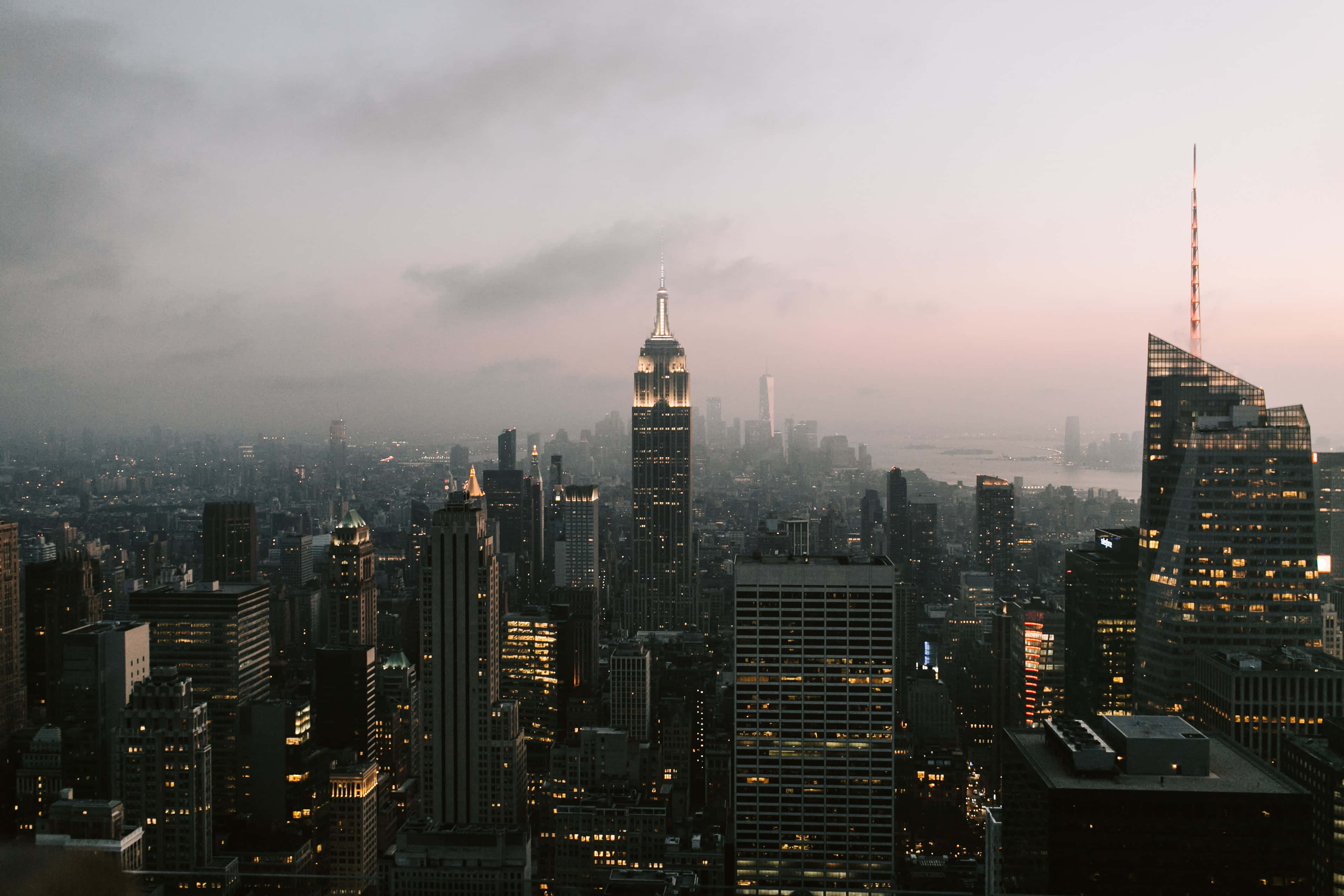 Nyc 桌布 壁紙 — 紐約市天際線的薄霧黃昏景觀，摩天大樓燈火通明，柔和的粉紅色和灰色雲彩。