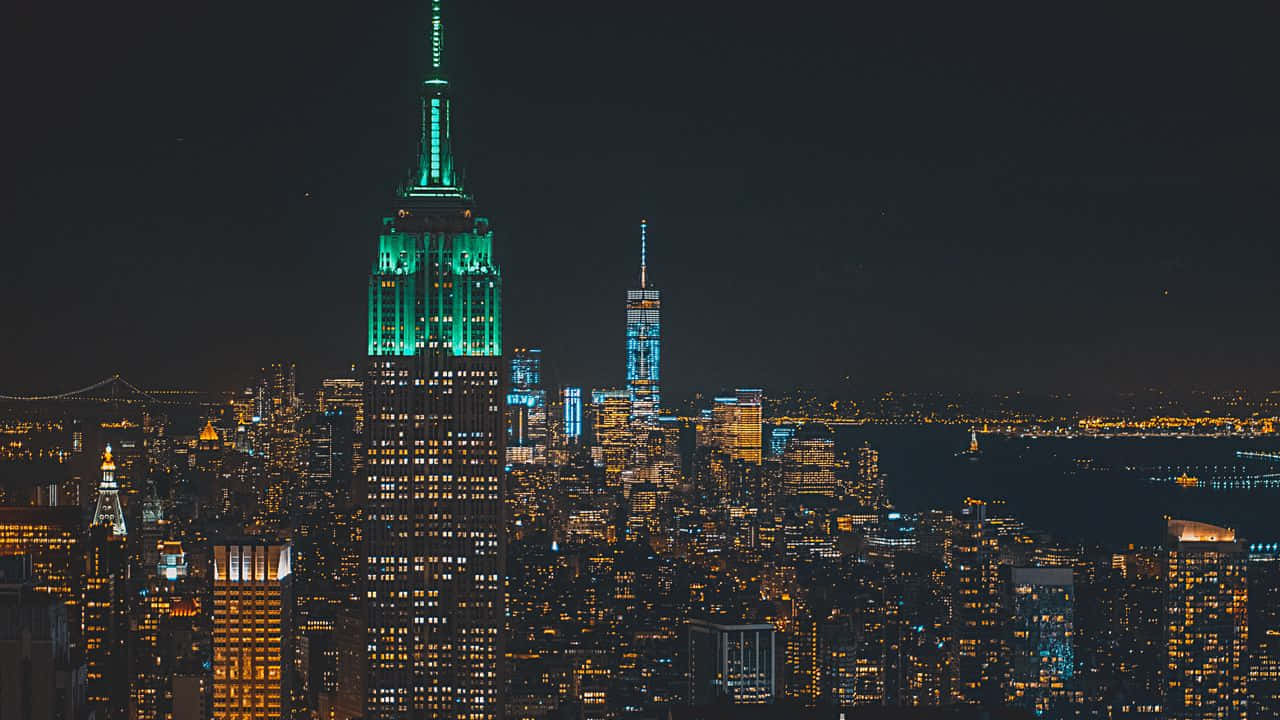 720p Empire State Building 桌布 壁紙 — 紐約市天際線的迷人夜景，帝國大廈閃爍著綠色光芒，周圍的摩天大樓則被溫暖的琥珀色燈光照亮。