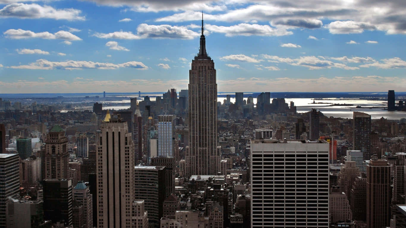 1366x768 Empire State Building 桌布 壁紙 — 紐約市天際線的鳥瞰圖，以帝國大廈為主導，在明亮的藍天和零星雲朵下。