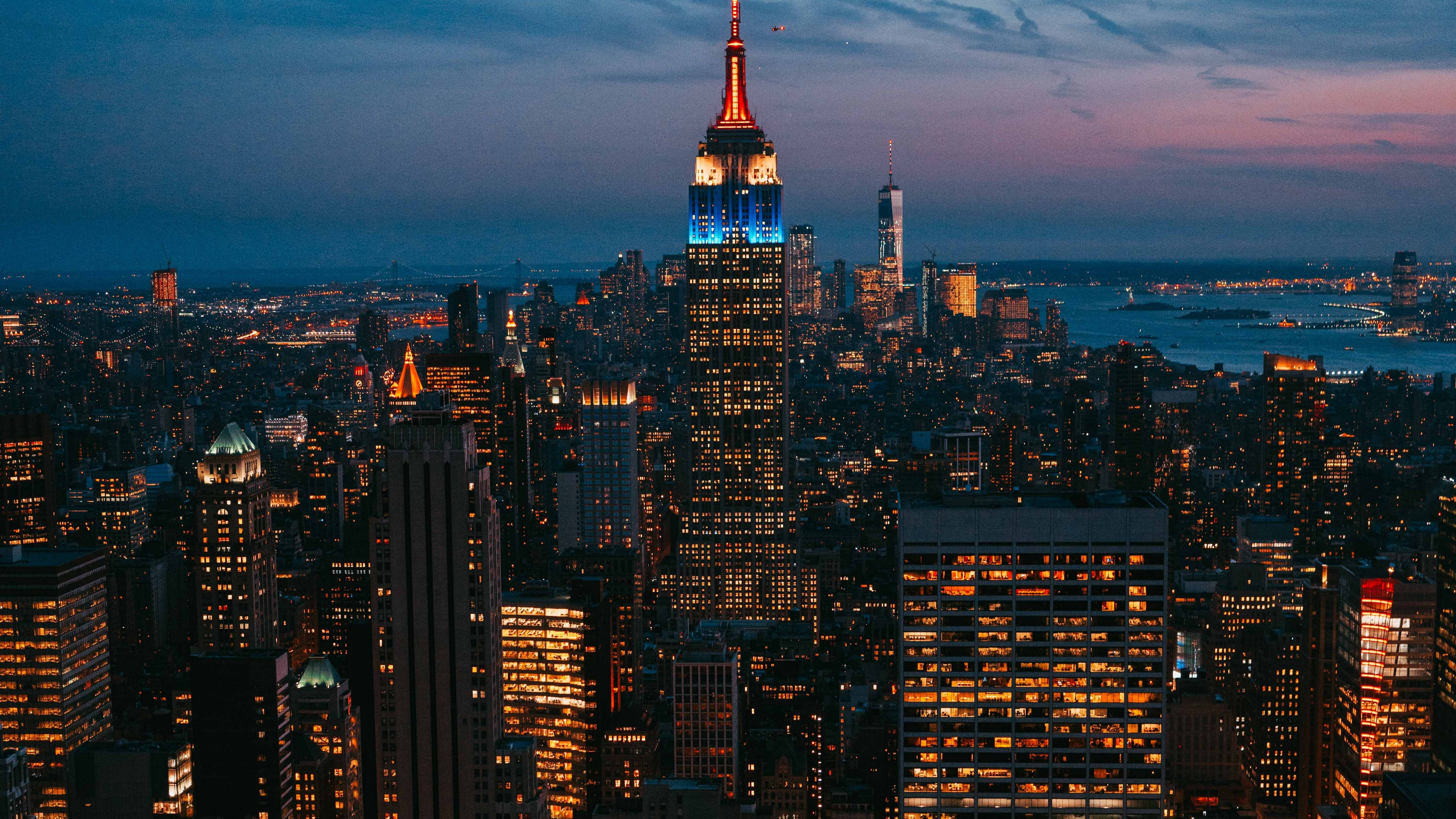 New York 桌布 壁紙 — 紐約市天際線的鳥瞰圖，在黃昏時分，帝國大廈在紫色天空下閃耀著藍色和橙色光芒。