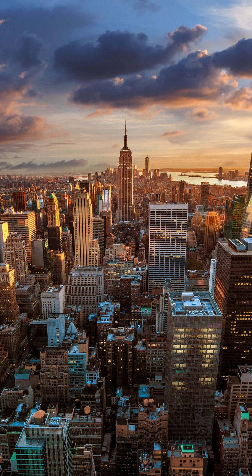 new york city iPhone x 桌布 壁紙 — 紐約市天際線的鳥瞰圖，日落時金色光芒照亮摩天大樓和上方戲劇性的雲層。