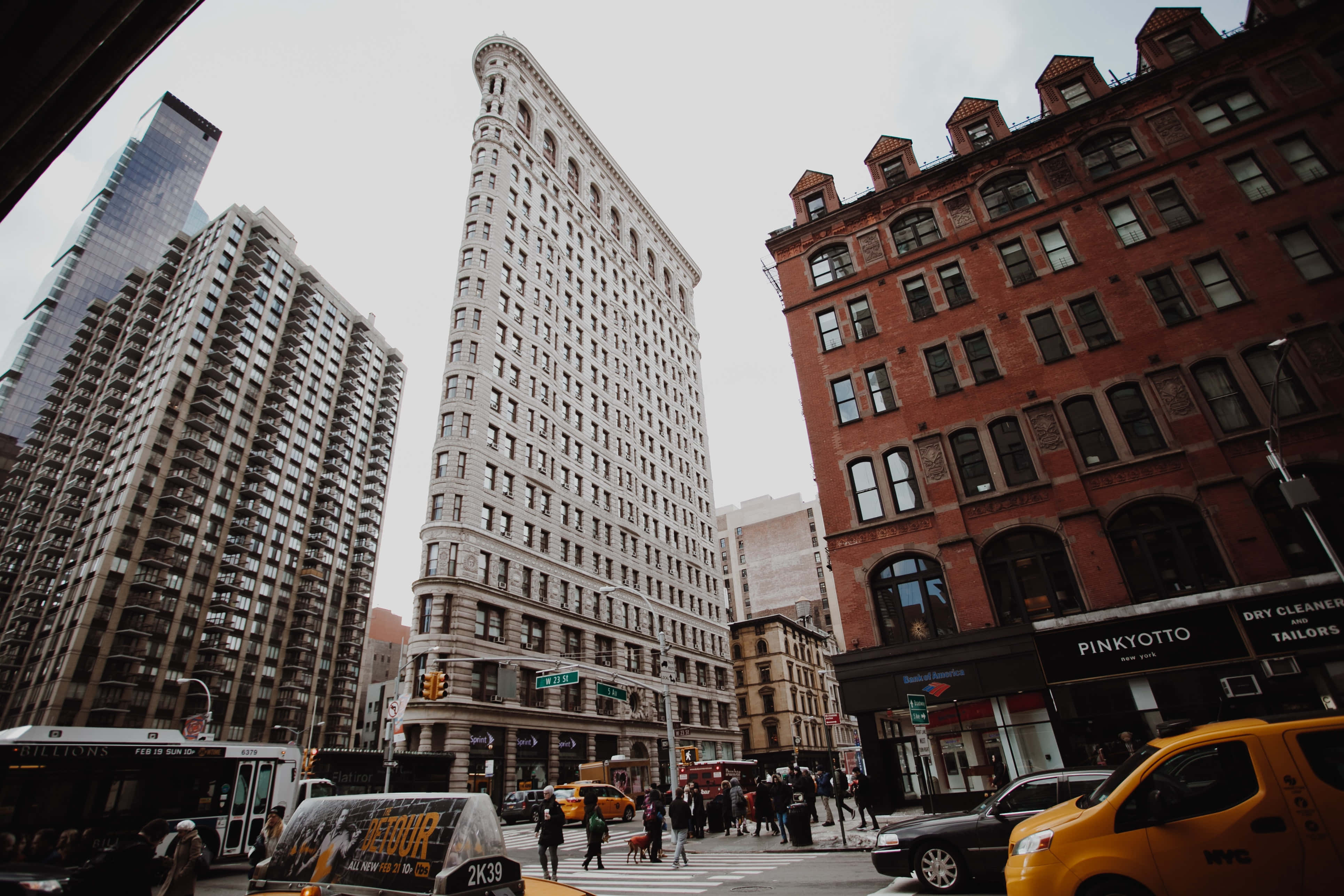 Nyc 桌布 壁紙 — 紐約市標誌性熨斗大廈的低角度景觀，聳立在紅磚和玻璃摩天大樓之間，下方有黃色計程車和行人。