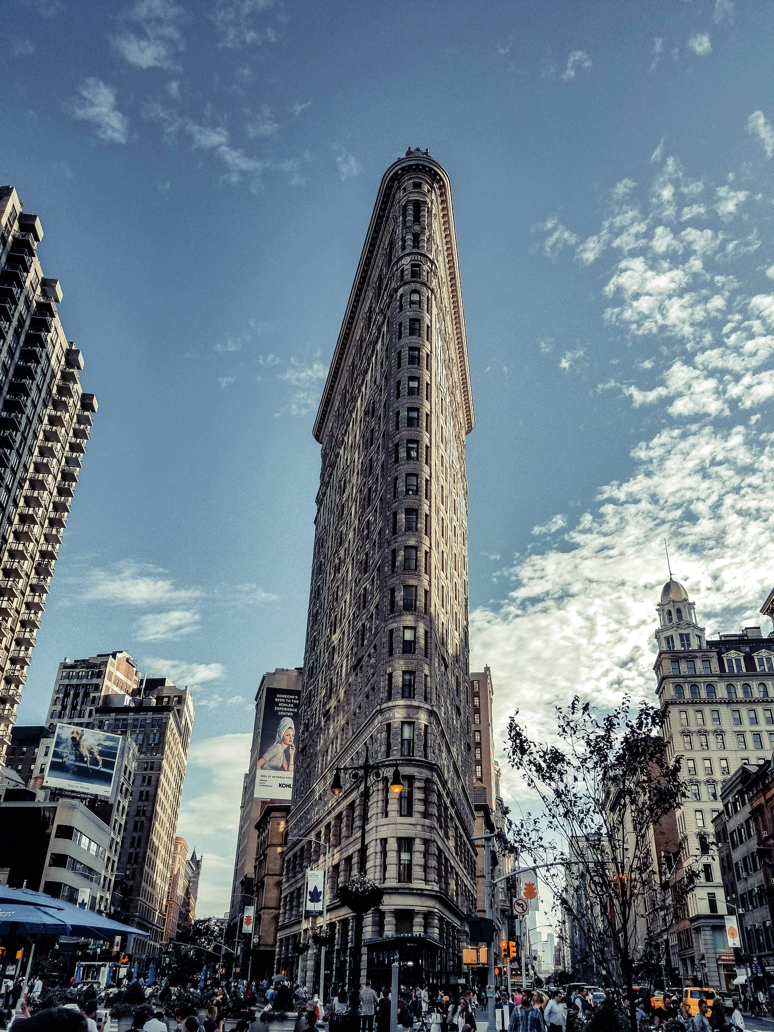 Flatiron Building 桌布 壁紙 — 紐約市標誌性的熨斗大廈在明亮的藍天和白雲下高聳入雲，周圍是繁忙的街道和行人。