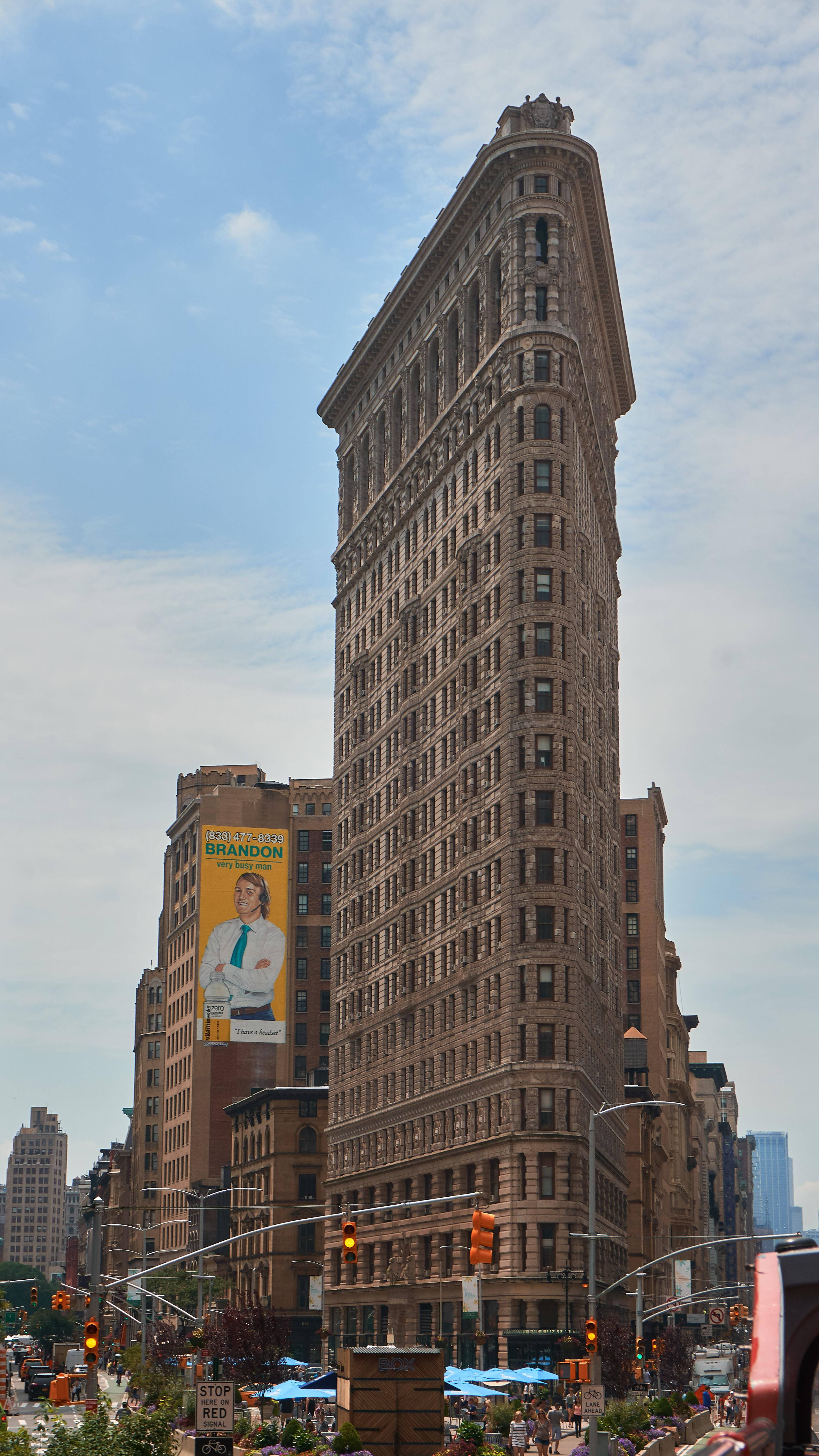 Flatiron Building 桌布 壁紙 — 紐約市標誌性的熨斗大廈高聳入雲，天空部分多雲，下方街道繁忙，行人如織。