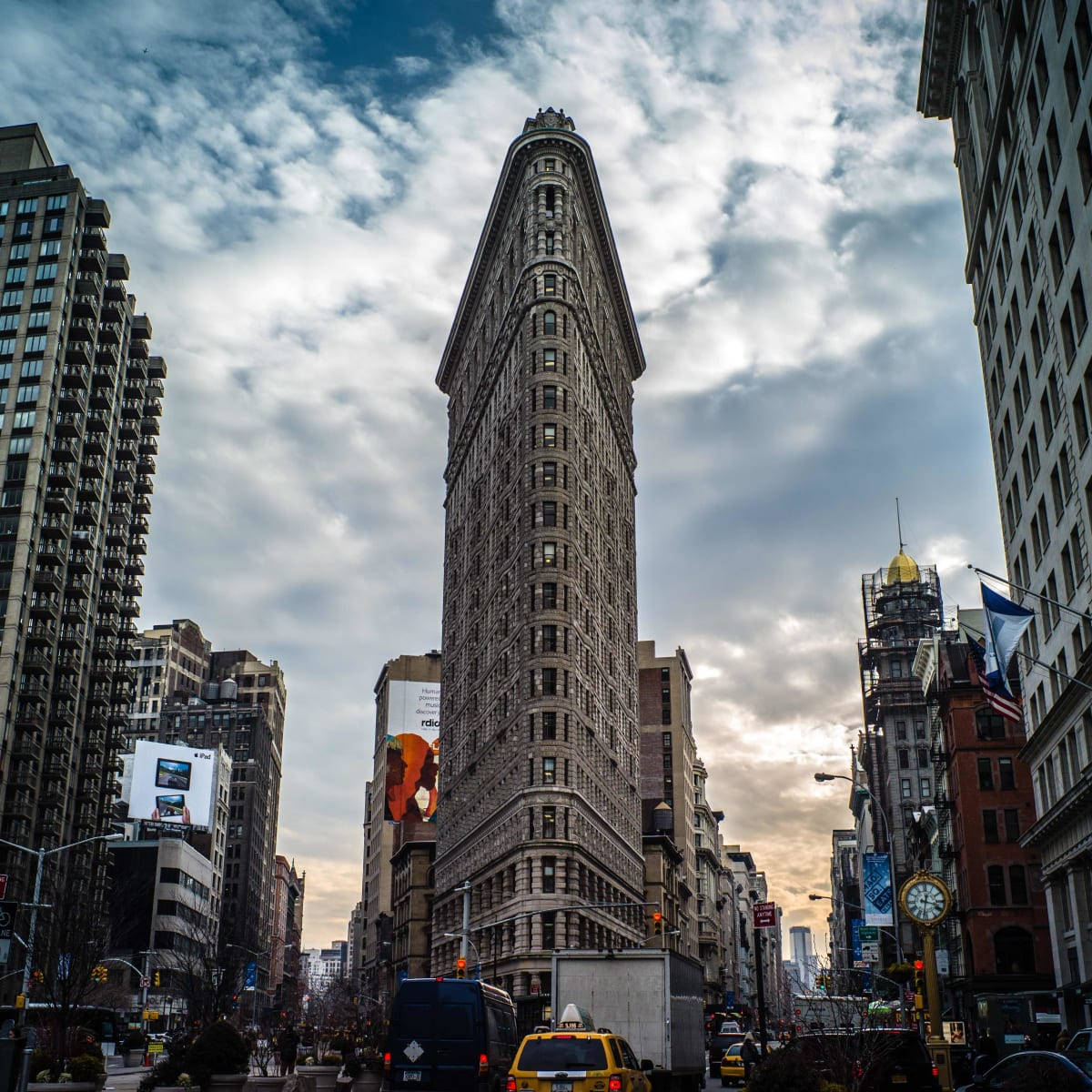 Flatiron Building 桌布 壁紙 — 紐約市的熨斗大廈在戲劇性的多雲天空下，聳立在繁華的城市街道上。