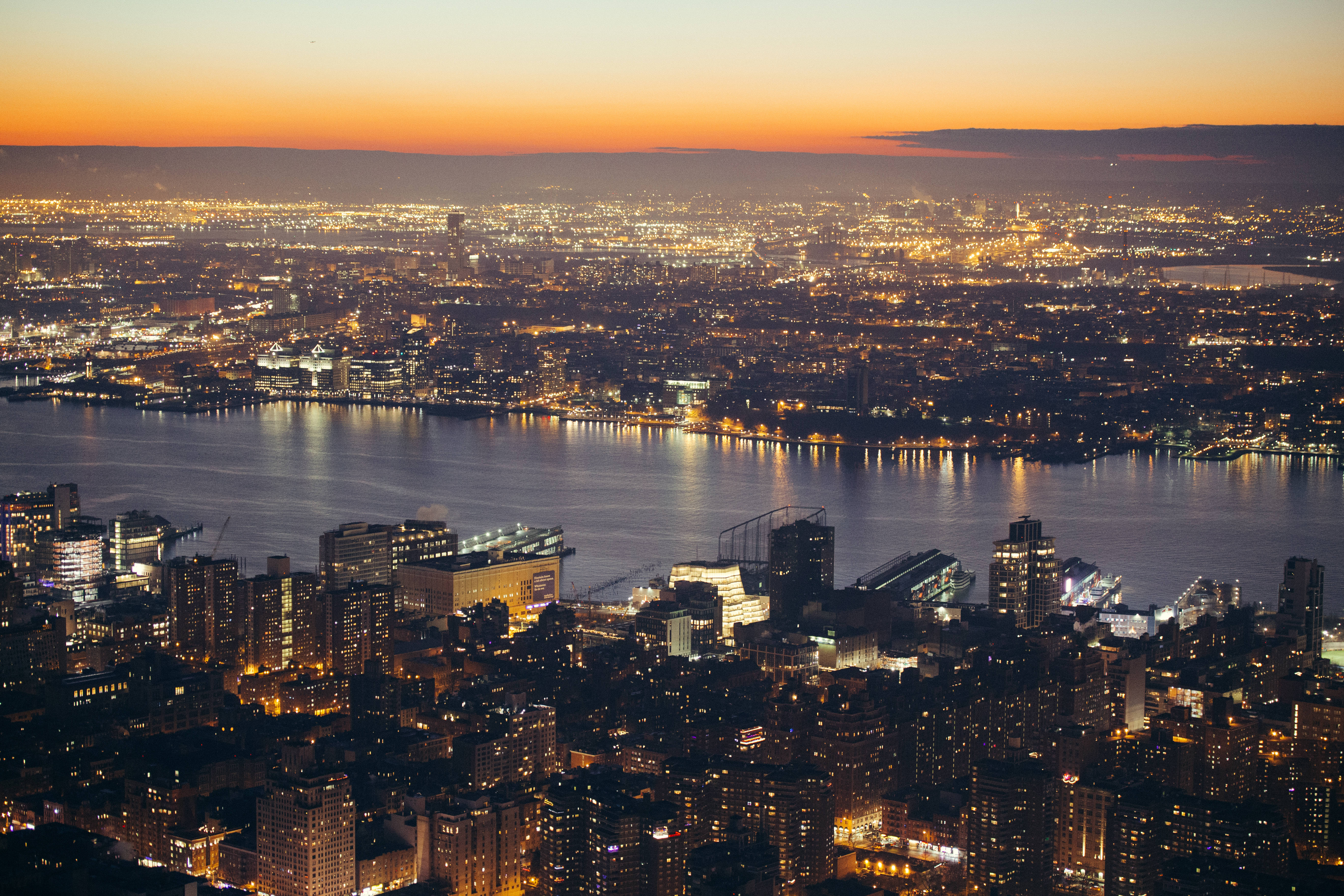 Nyc 桌布 壁紙 — 紐約市黃昏時的鳥瞰圖，閃爍的摩天大樓和一條河流在橙色天空下反射城市燈光。