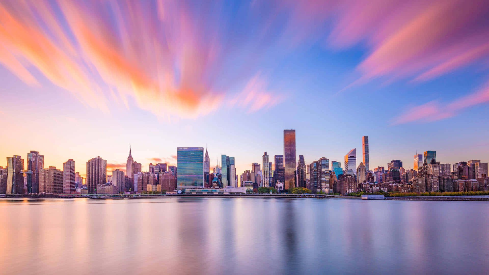 New York Art 桌布 壁紙 — 紐約日落時分的長時間曝光城市景觀，平靜的水面倒映著粉紅、橙和藍色的條紋雲彩。