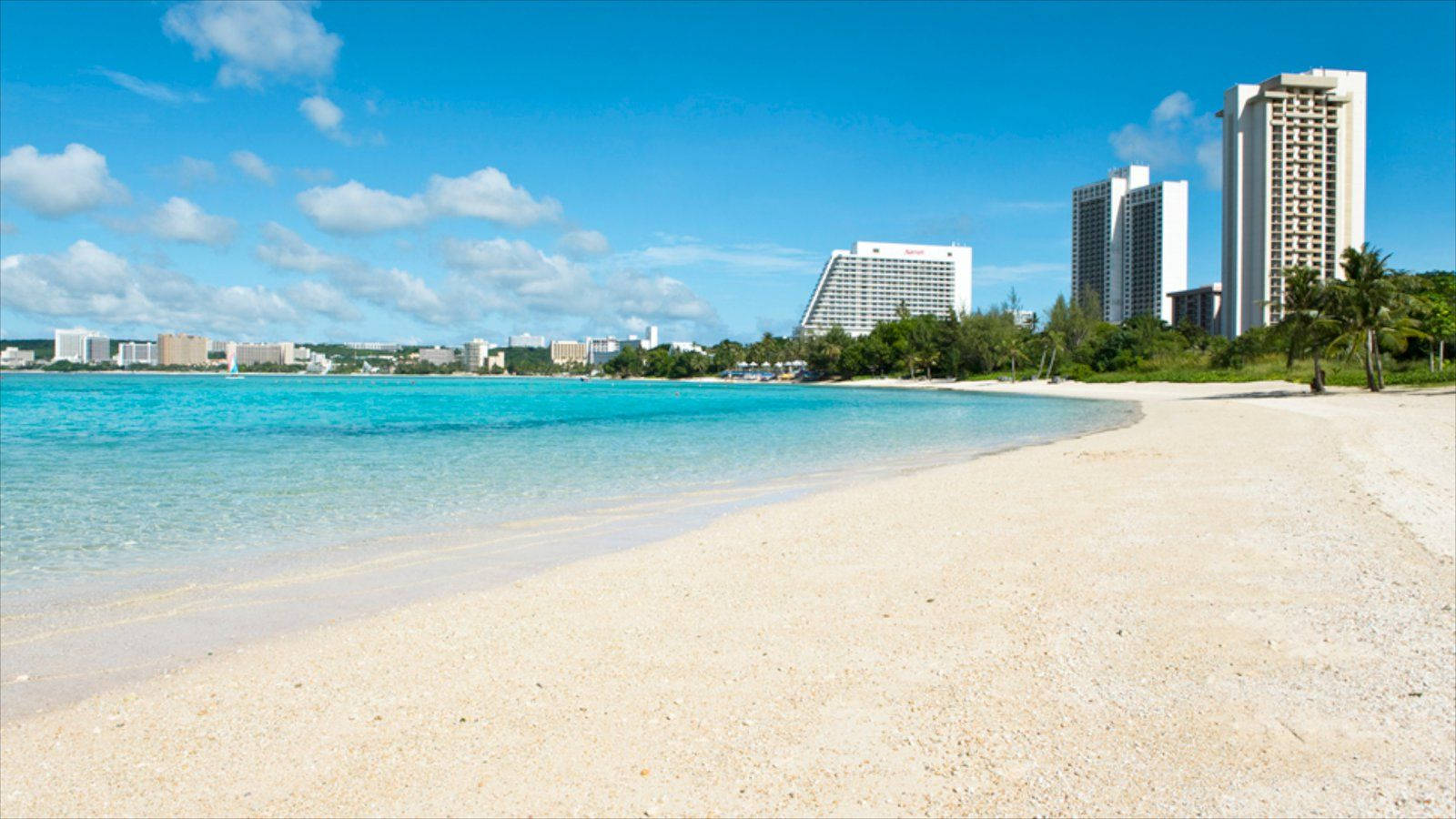 Guam 桌布 壁紙 — 純淨的沙灘沿著碧綠的海水蜿蜒伸展，現代化的酒店在茂密的綠植後方聳立，頭頂是明亮的藍天。