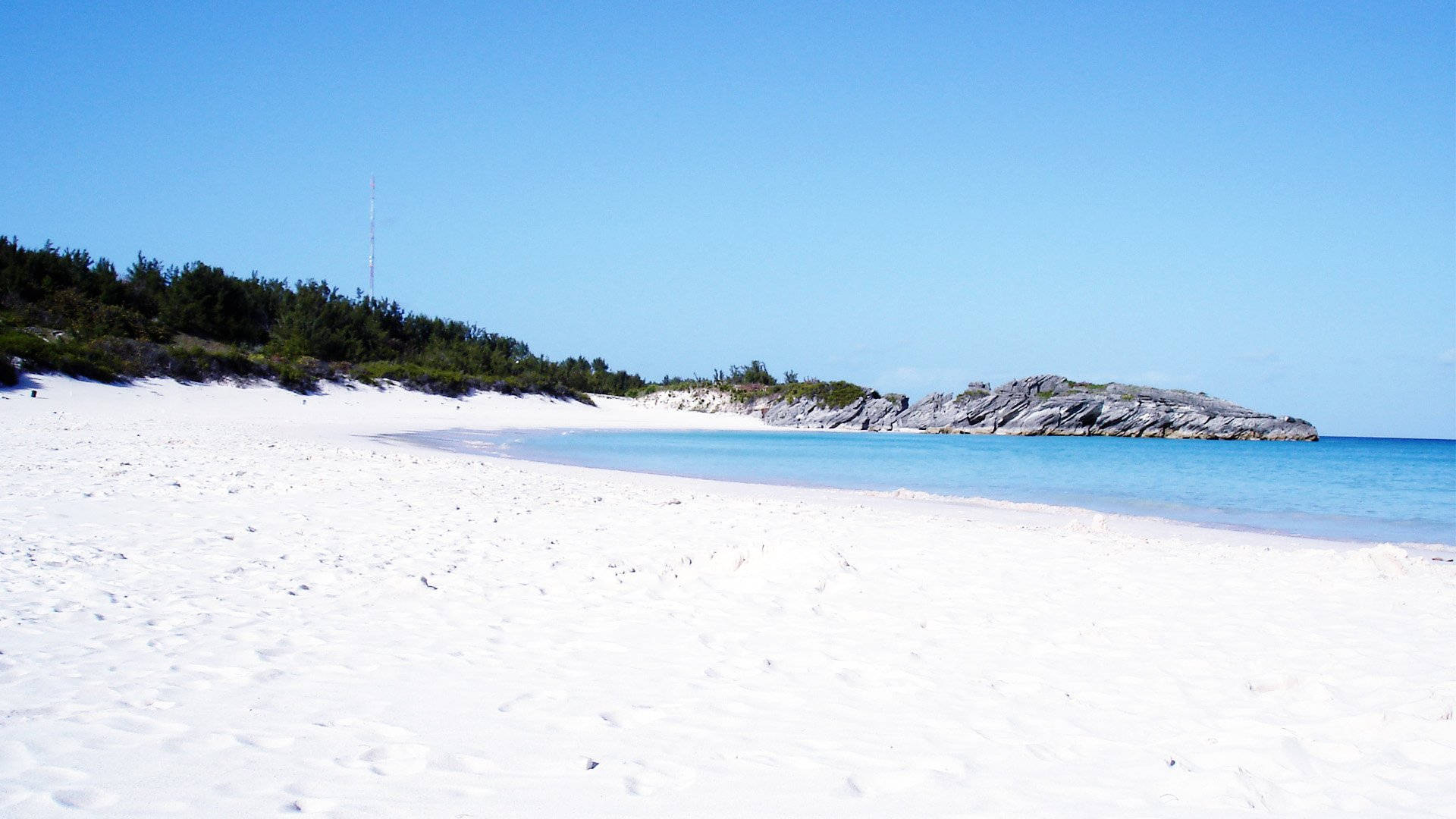 Bermuda 桌布 壁紙 — 純淨的白色沙灘在清澈的藍天下緩緩彎向碧綠的海水，遠處有岩石露頭和綠色灌木。