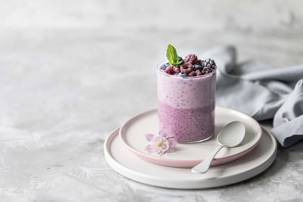 Cute Dessert 桌布 壁紙 — 紫色分層奇亞籽布丁，上面放有莓果和薄荷，放在粉彩盤子上，旁邊有精緻的花朵和湯匙。