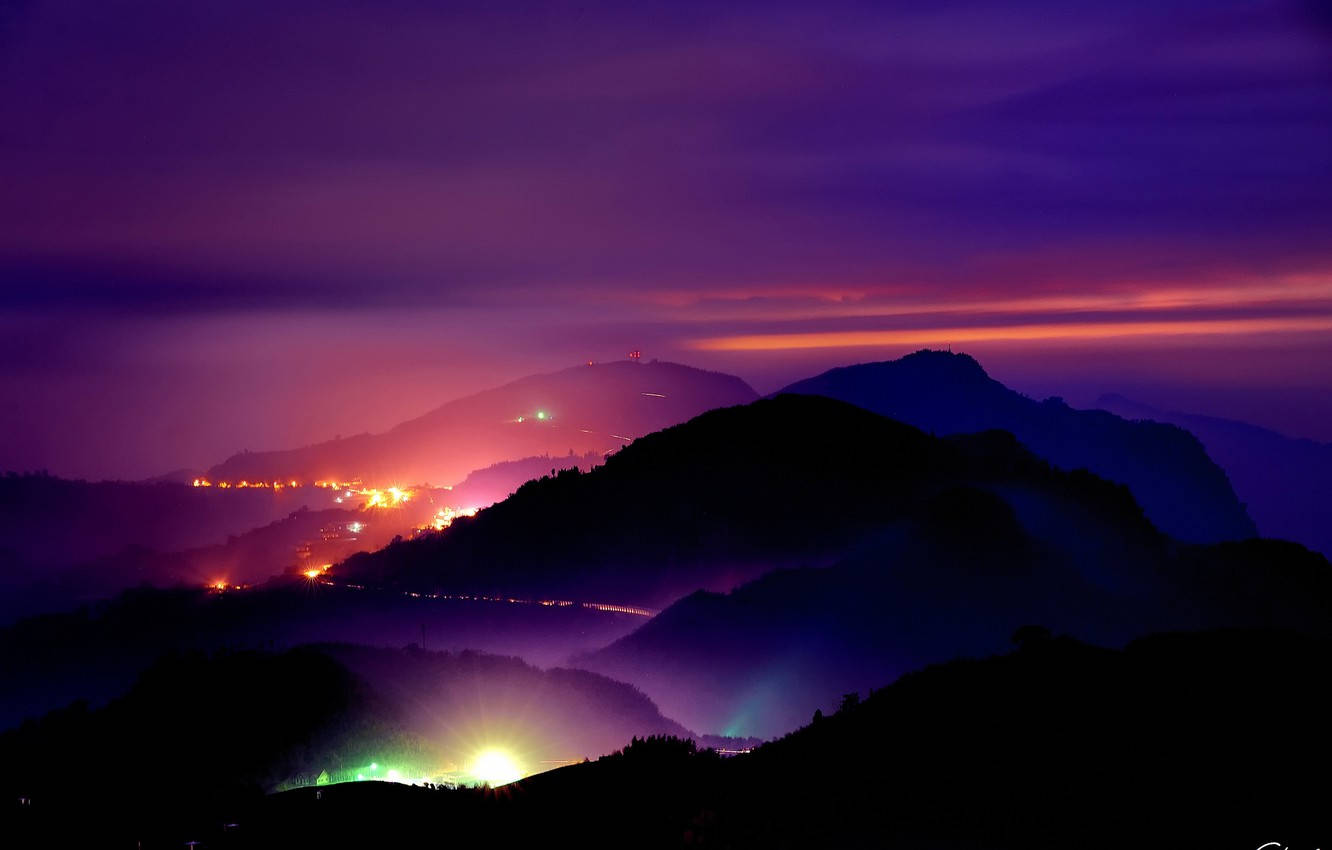 View 桌布 壁紙 — 紫色和橙色的暮色覆蓋著層層疊疊的山脊，山谷中村莊燈火閃爍。
