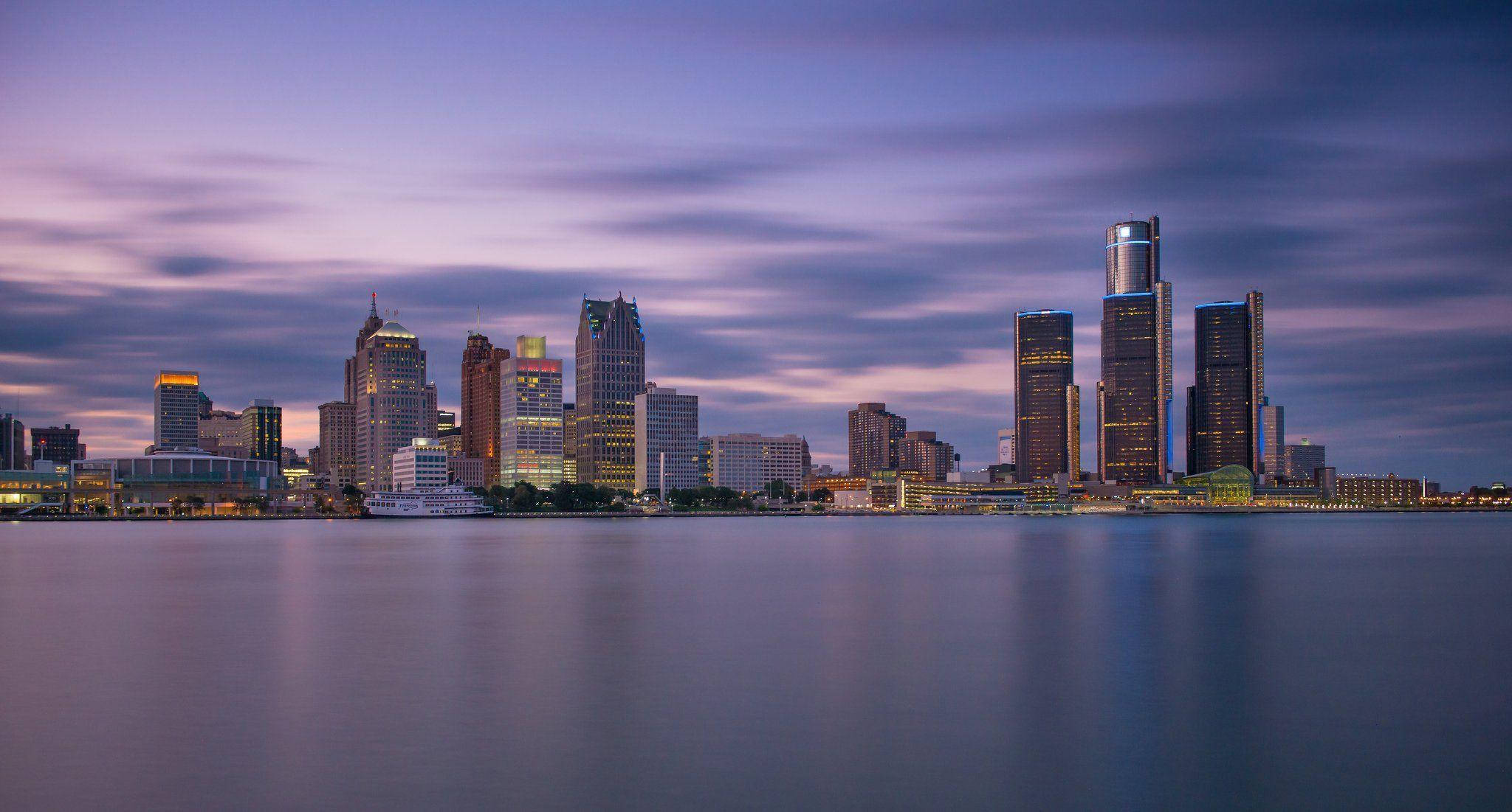 detroit 桌布 壁紙 — 紫色天空下，高樓大廈倒映在平靜水面的黃昏城市天際線，氣氛寧靜。