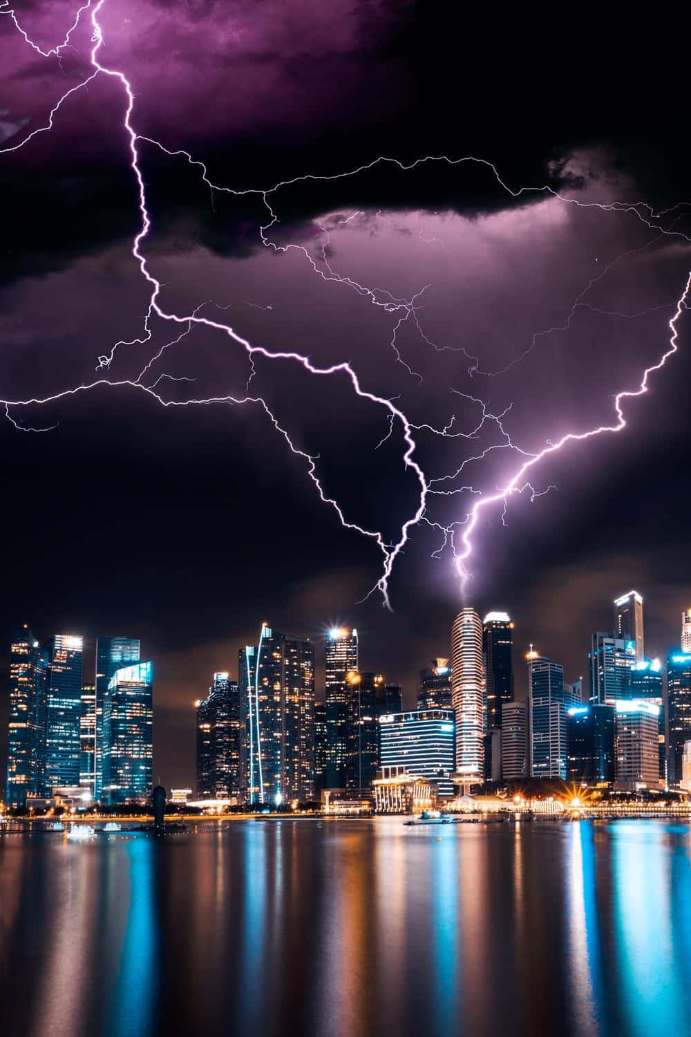 Lightning Bolt 桌布 壁紙 — 紫色閃電在暴風雨的夜空中分叉，映照在平靜水面上的發光城市天際線之上。
