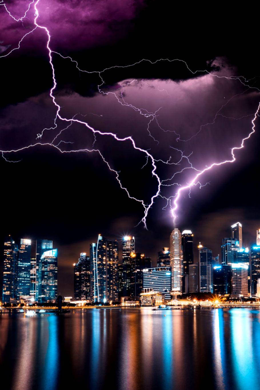 閃電 桌布 壁紙 — 紫色閃電在暴風雨的天空中分叉，映照在黑暗水面上發光的城市天際線之上。