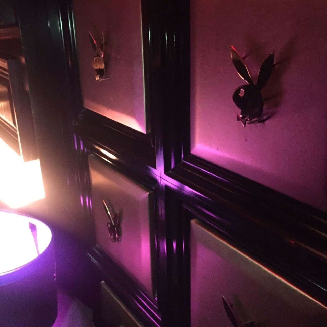 Playboy Aesthetic 桌布 壁紙 — 紫色霓虹燈照亮了黑色框架的面板，上面裝飾著金屬質感的Playboy兔子標誌，室內光線昏暗。