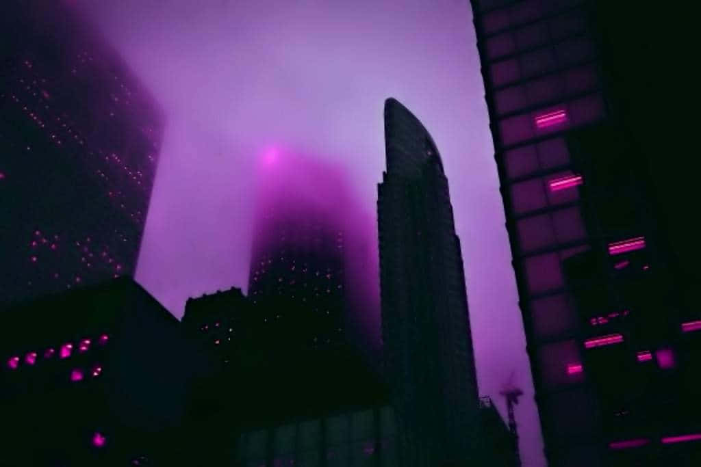 城市 桌布 壁紙 — 紫色霓虹燈穿過朦朧的夜空，照亮高聳的摩天大樓，呈現未來主義的城市景觀。