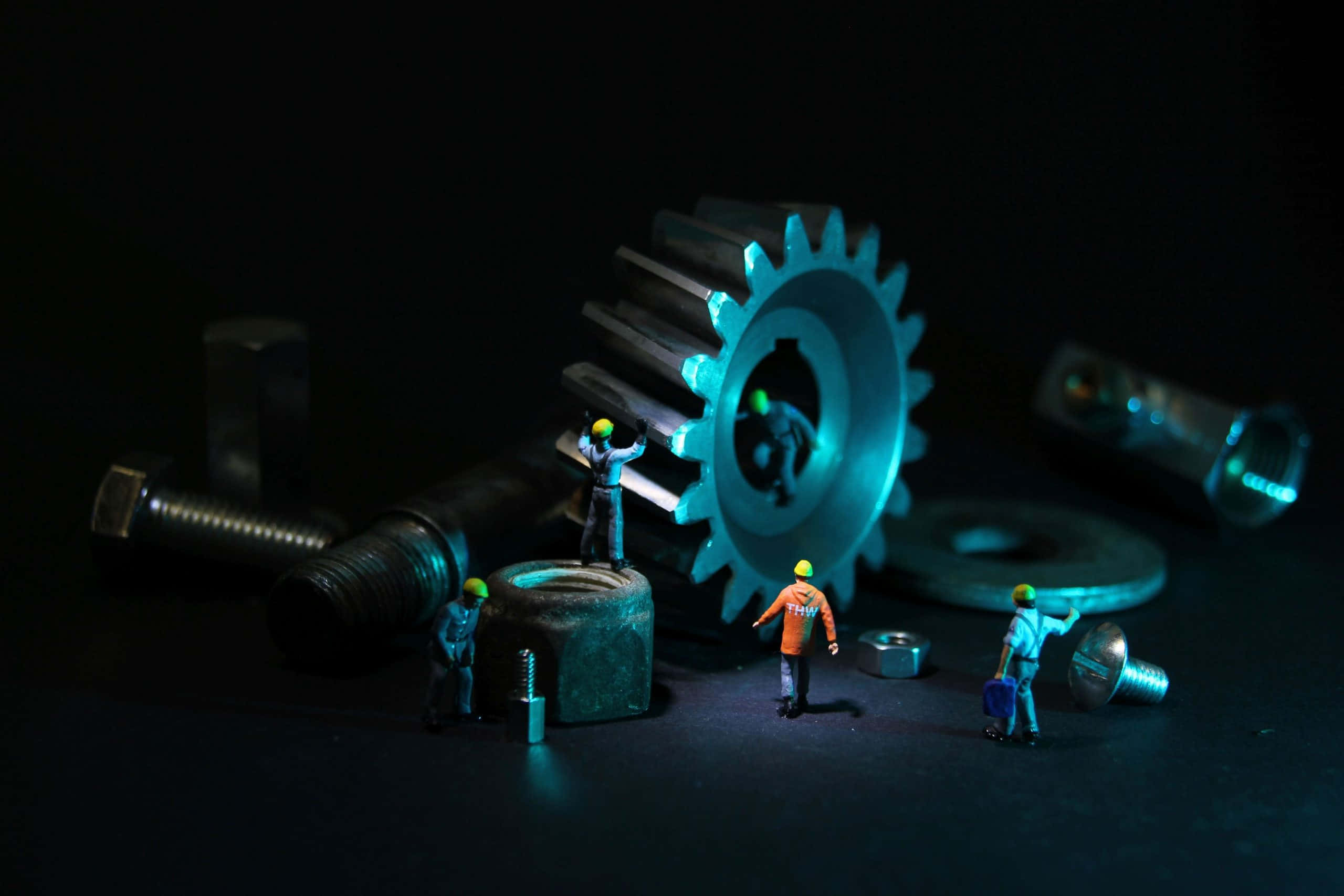 engineering 桌布 壁紙 — 細小的建築工人戴著頭盔，在黑暗背景前組裝發光的藍色齒輪和螺栓。