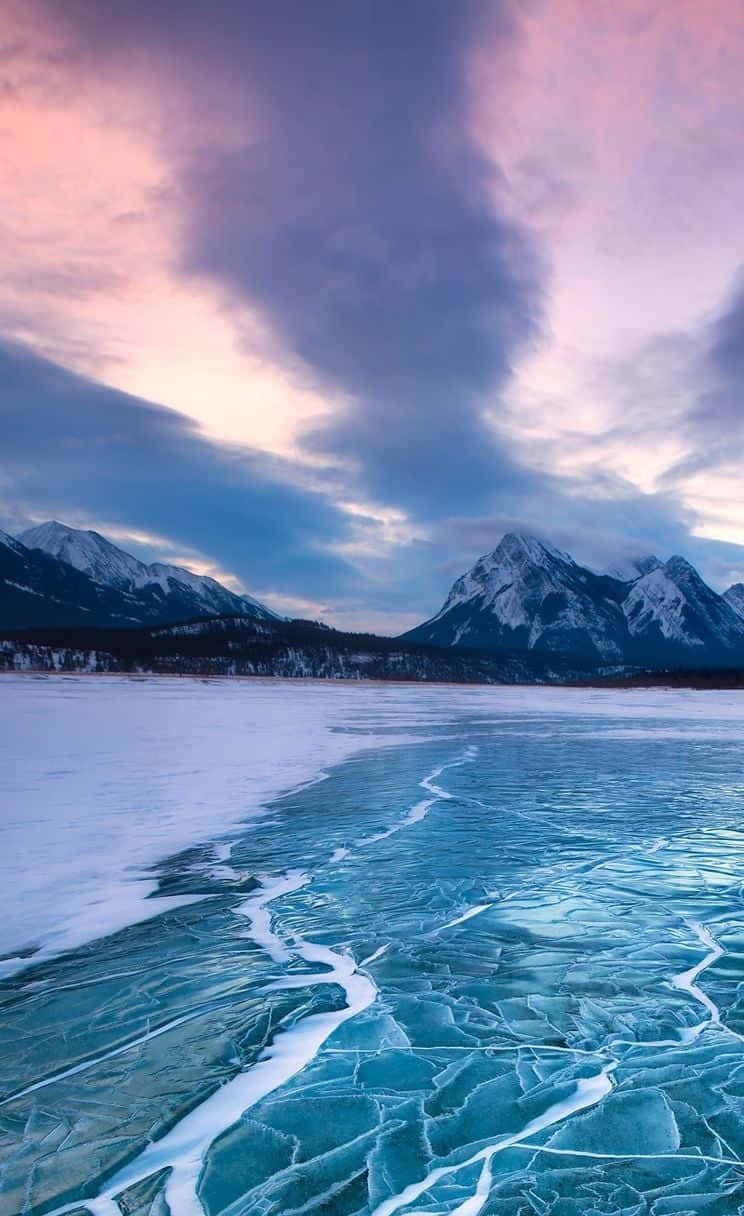 Frozen Lake 桌布 壁紙 — 結冰的湖面佈滿裂紋的綠松石冰塊，在戲劇性的粉紫暮色天空下，與白雪覆蓋的湖岸相接，遠方是積雪的山峰。