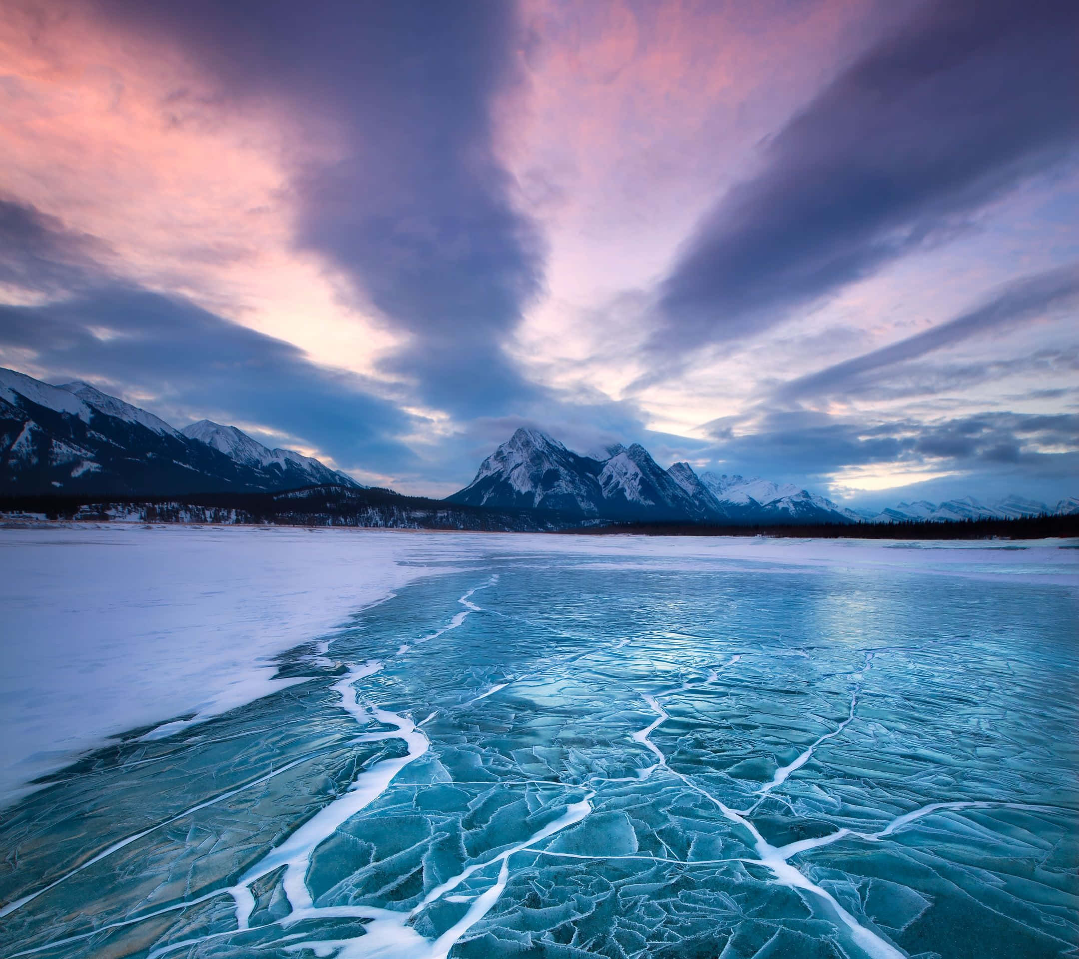 Frozen Lake 桌布 壁紙 — 結冰的湖面帶著裂縫的藍色冰塊，在戲劇性的粉紅和紫色日落天空下，與積雪覆蓋的山脈相遇。