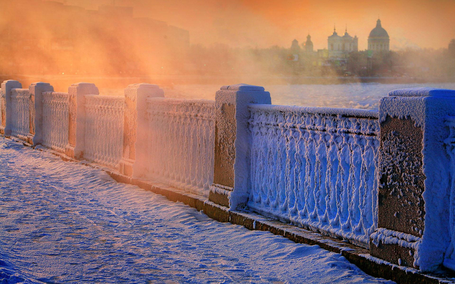 Saint Petersburg 桌布 壁紙 — 結霜的橋樑欄杆在溫暖的日出下閃耀，霧氣從結冰的河面升起，遠處有圓頂建築。