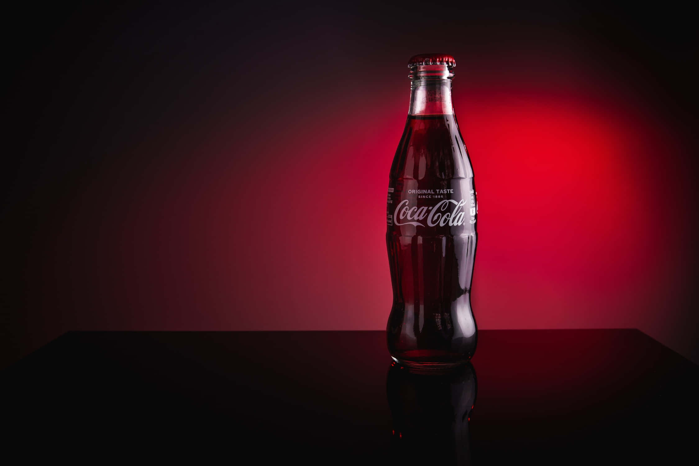 coca cola 桌布 壁紙 — 經典可口可樂玻璃瓶在深紅色漸變背景和戲劇性工作室燈光下發光。