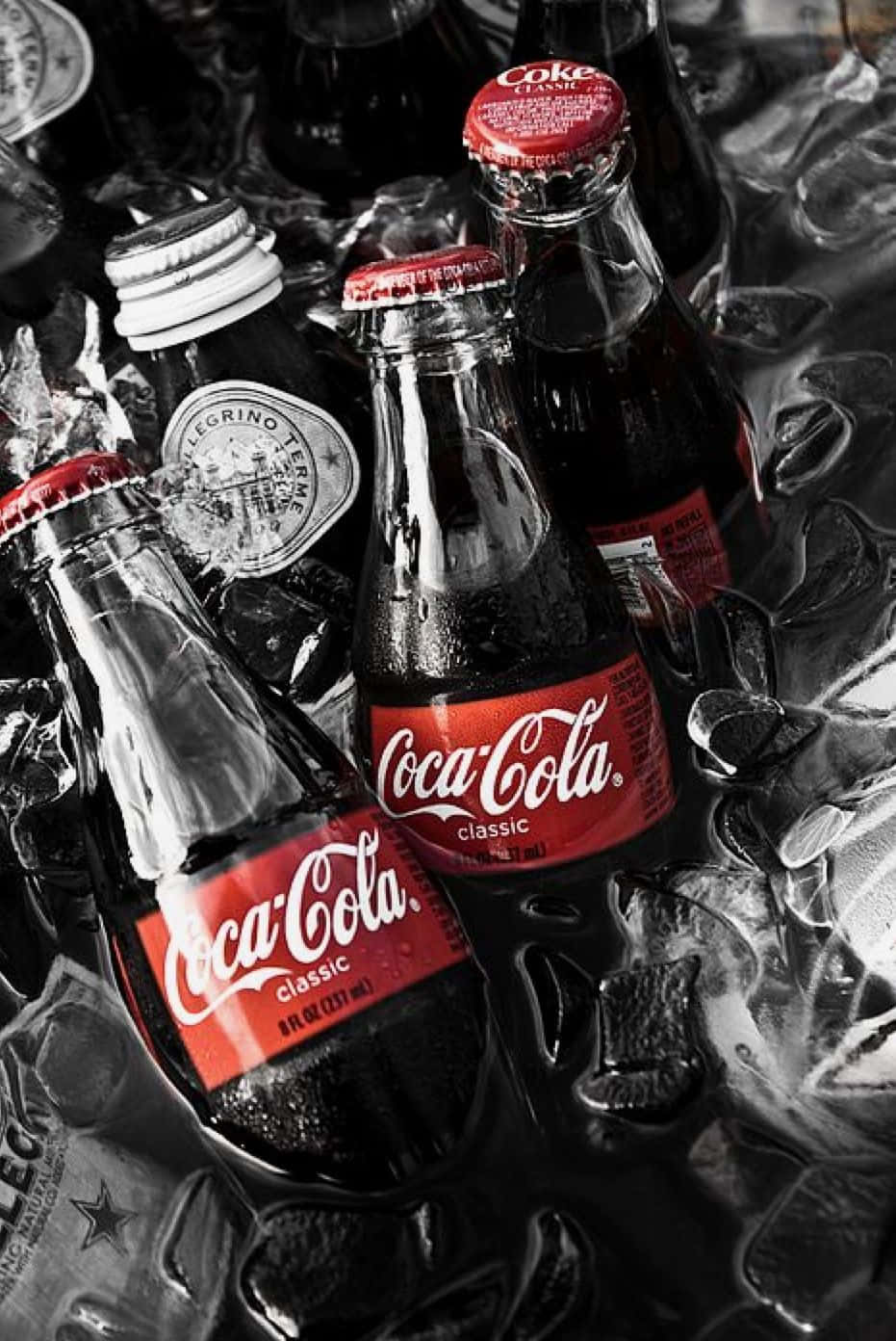 coca cola 桌布 壁紙 — 經典可口可樂瓶帶紅色標籤，冰凍在閃閃發光的冰塊中，單色構圖帶有選擇性紅色。