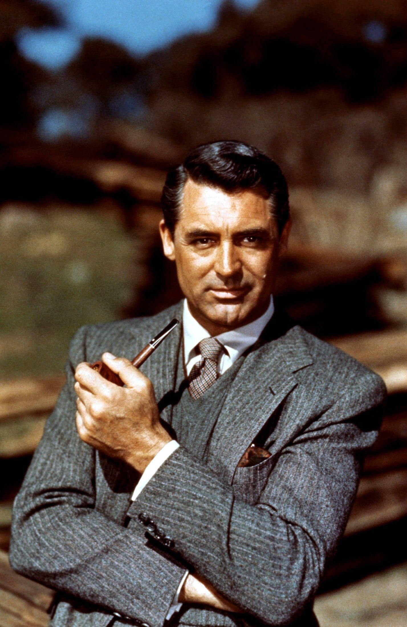 Celebrity 桌布 壁紙 — 經典好萊塢演員 Cary Grant 身穿筆挺灰色西裝，手持煙斗，自信地凝視著陽光普照的戶外背景。