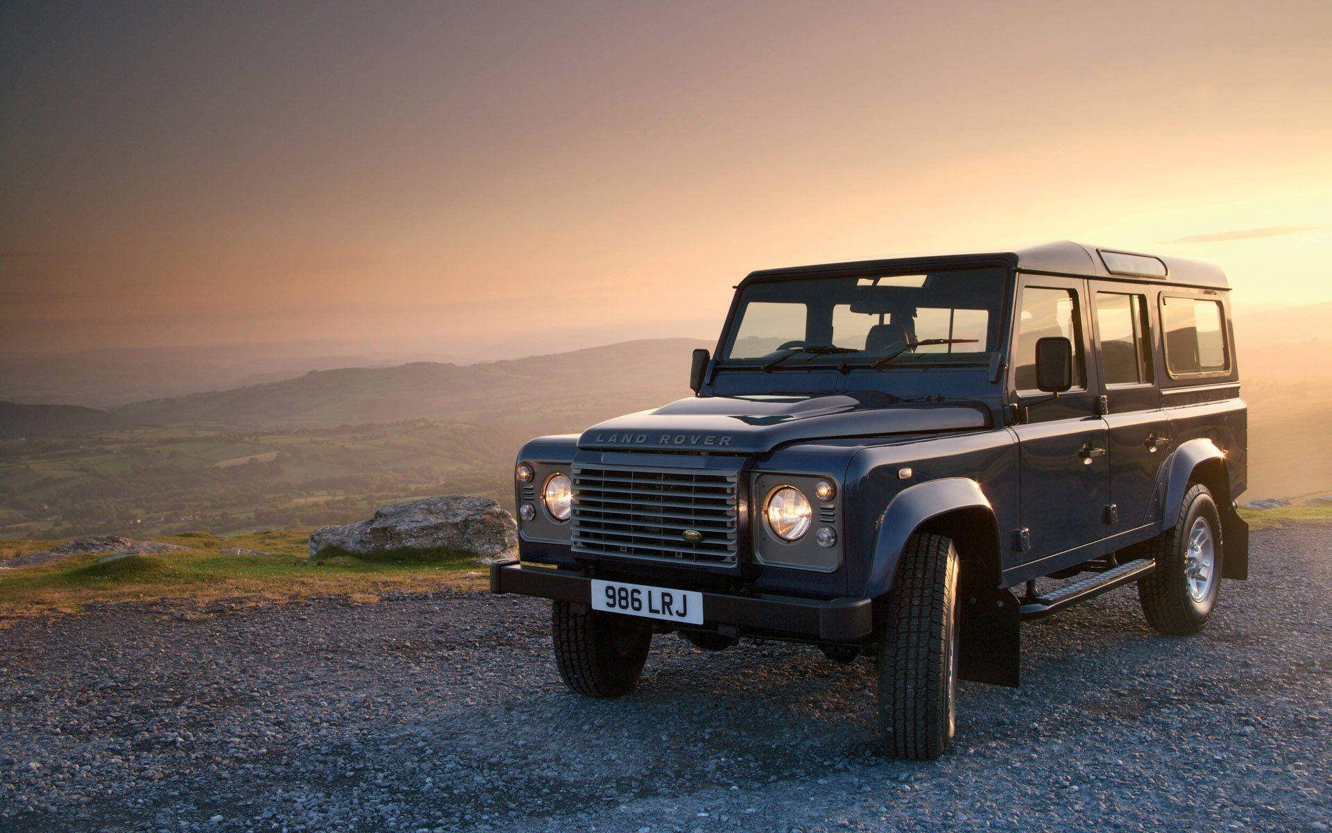 荒原路華 桌布 壁紙 — 經典深藍色 Land Rover Defender 停泊在碎石山頂，金色日落映襯著連綿起伏的山丘。