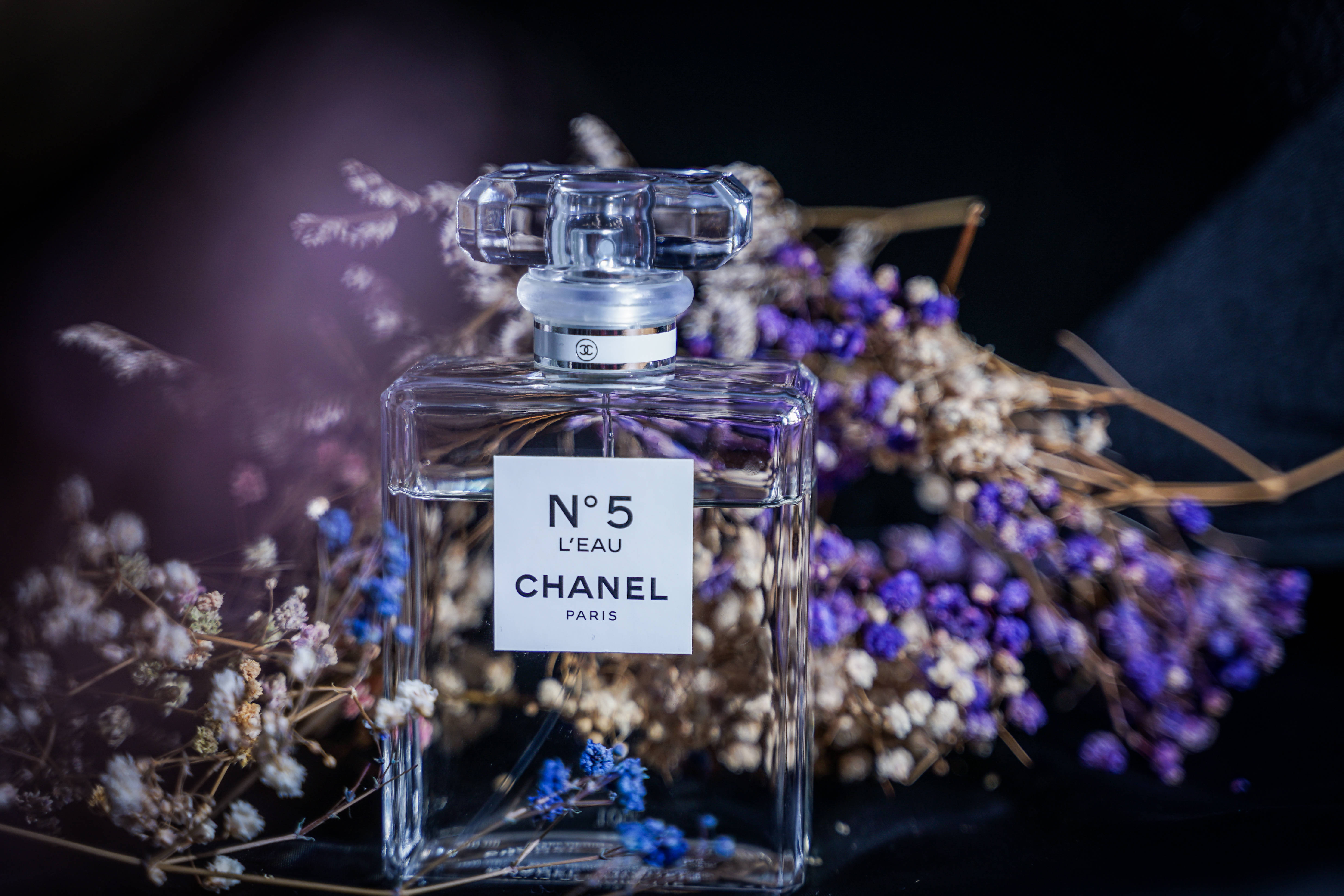 chanel iman 桌布 壁紙 — 經典的 Chanel No 5 香水瓶在柔和光線下閃耀，置於精緻的乾燥紫色和白色花朵之間，背景深色。