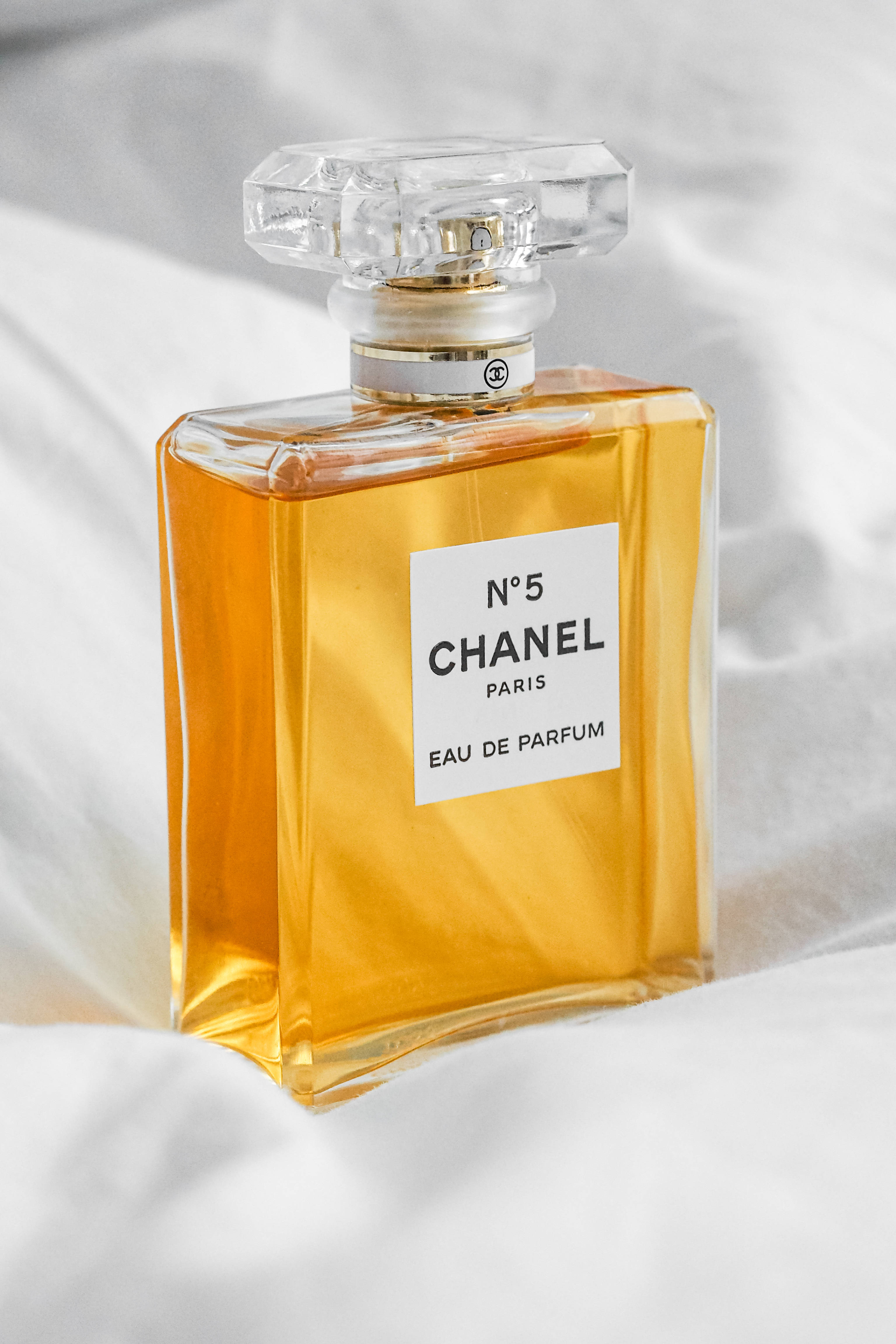 Chanel 桌布 壁紙 — 經典的Chanel No 5香水瓶裝有琥珀色液體，在明亮光線下優雅地放置在柔軟的白色布料上。