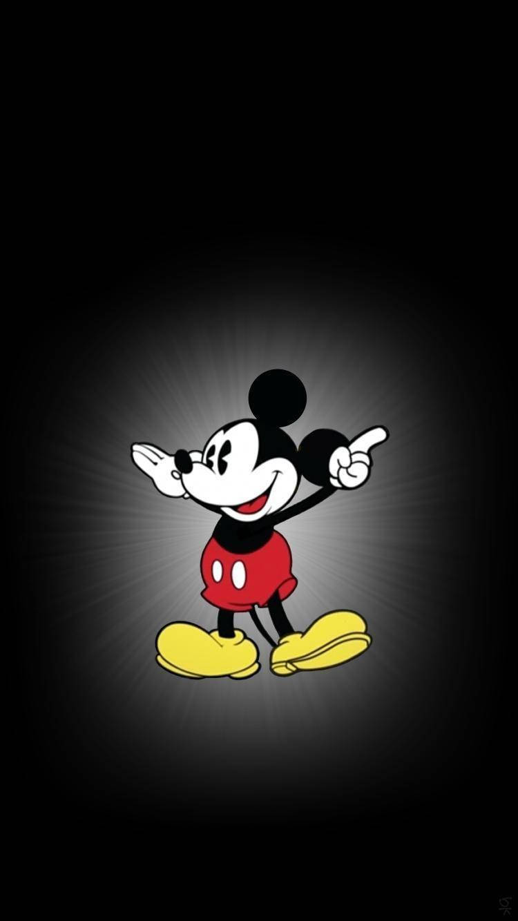 Mickey 桌布 壁紙 — 經典米奇老鼠穿著紅色短褲和黃色鞋子，在帶有聚光燈效果的深色背景前愉快地指著。