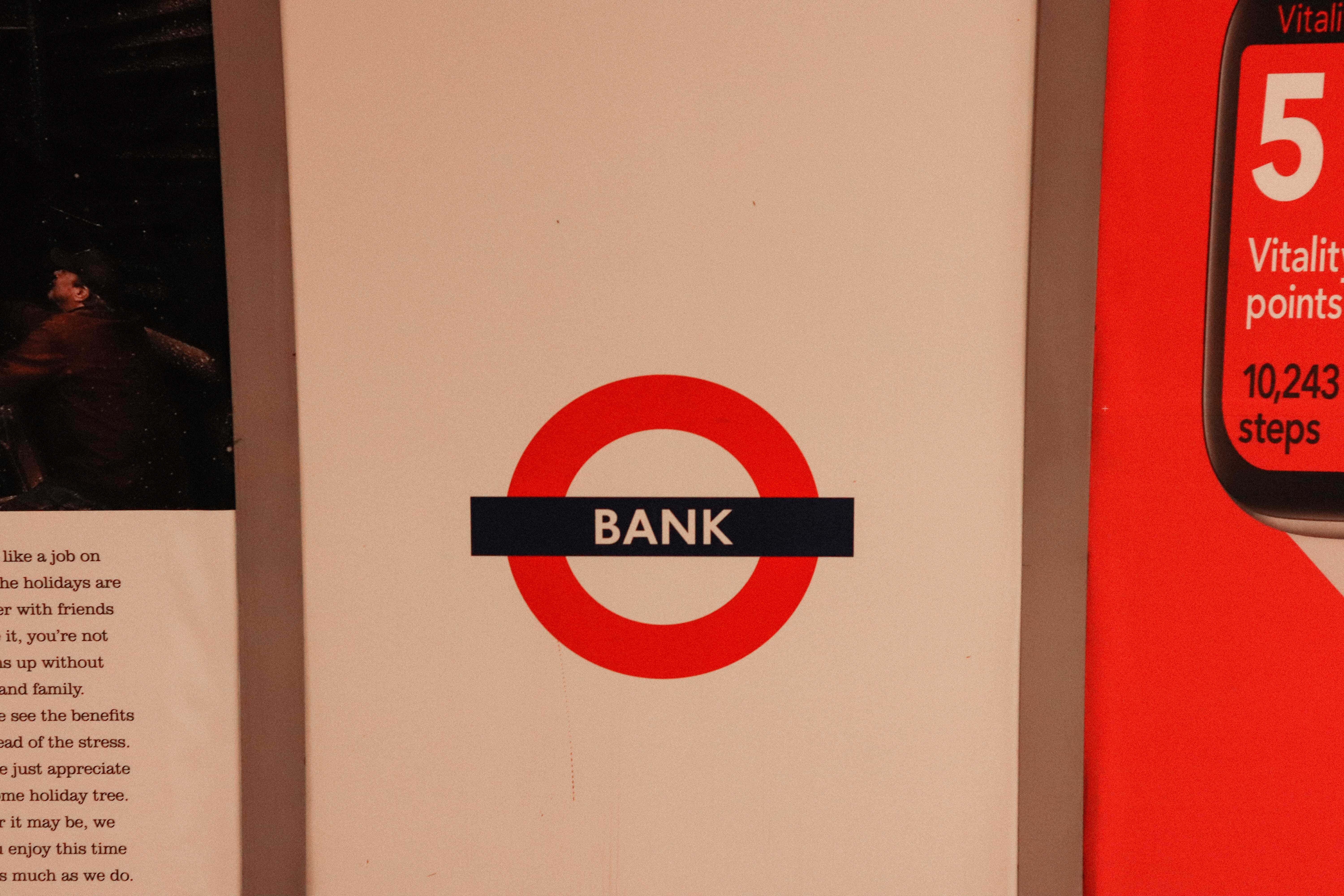 Bank 桌布 壁紙 — 經典紅色倫敦地鐵「BANK」車站標誌的特寫，位於米色牆壁上，旁邊有一個健身追蹤器顯示屏。