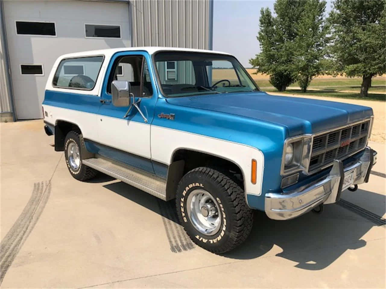 Gmc Jimmy 桌布 壁紙 — 經典藍白 Chevrolet K5 Blazer，帶有鍍鉻裝飾和堅固輪胎，在陽光明媚的日子裡停放在金屬車庫前。