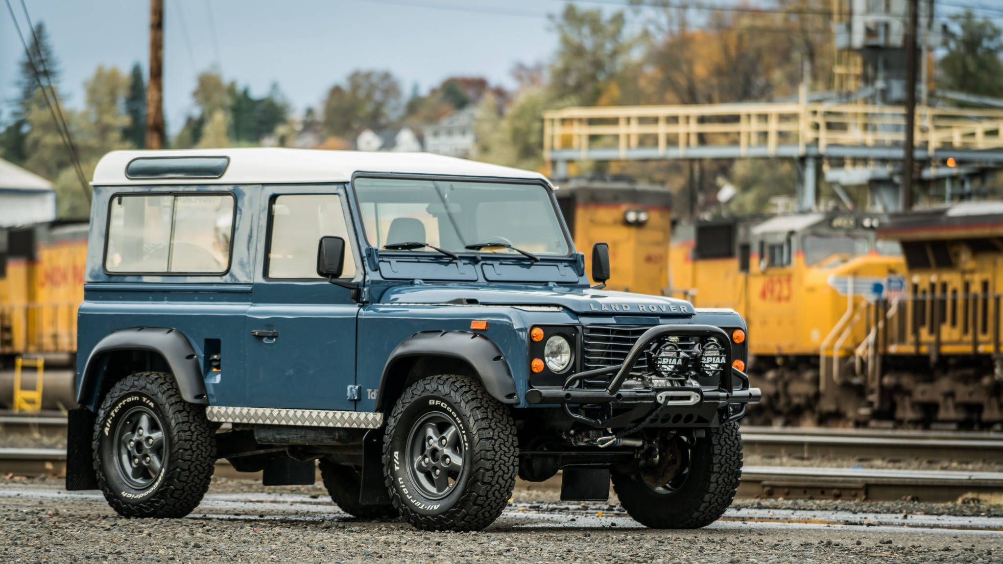 land rover iPhone 桌布 壁紙 — 經典藍色 Land Rover Defender 堅固地停在鐵軌旁的碎石上，背景有黃色火車頭。