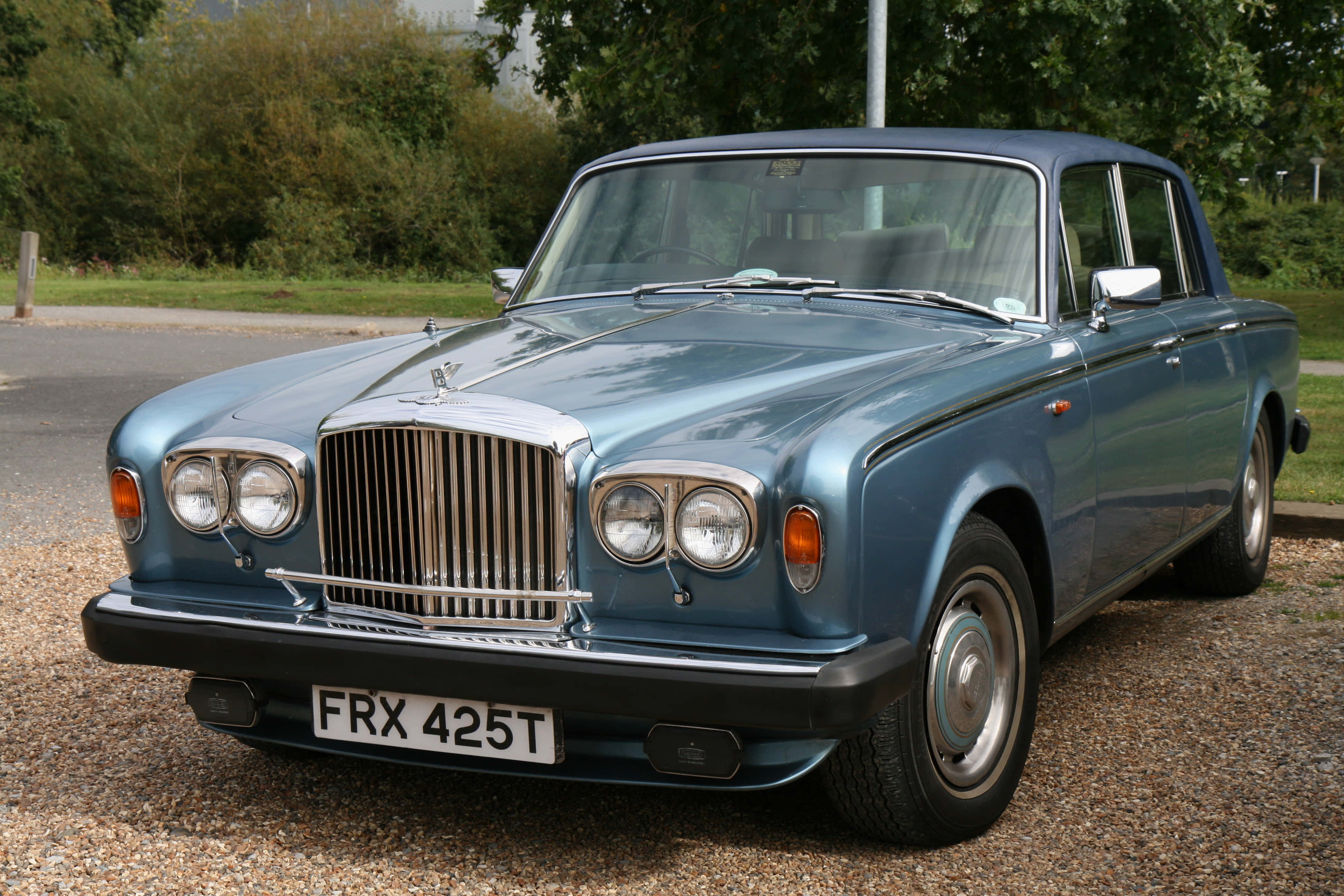 Bentley T2 桌布 壁紙 — 經典藍色賓利S3轎車在日光下閃耀，停泊在碎石上，旁邊有綠色樹葉。
