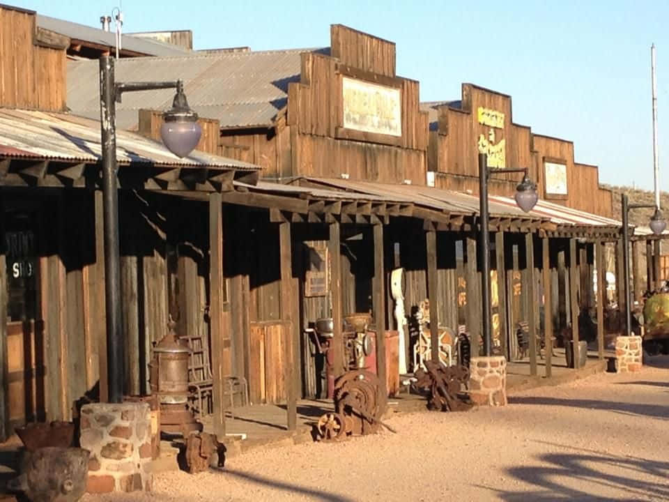 Old Western 桌布 壁紙 — 經典西部小鎮的 dusty 街道上，排列著陽光照射的木製店面，帶有生鏽的馬車輪和復古招牌。