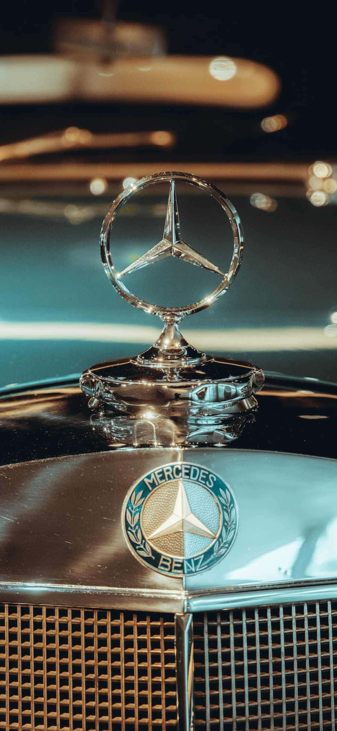 Mercedes 桌布 壁紙 — 經典賓士引擎蓋標誌特寫，在溫暖環境光下閃爍，背景反射模糊。