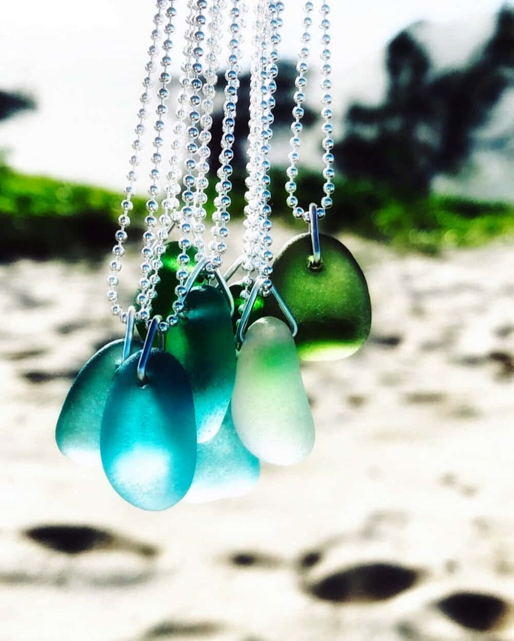 Sea Glass 桌布 壁紙 — 綠松石和綠色的海玻璃寶石掛在銀鏈上，背景是模糊的海灘。