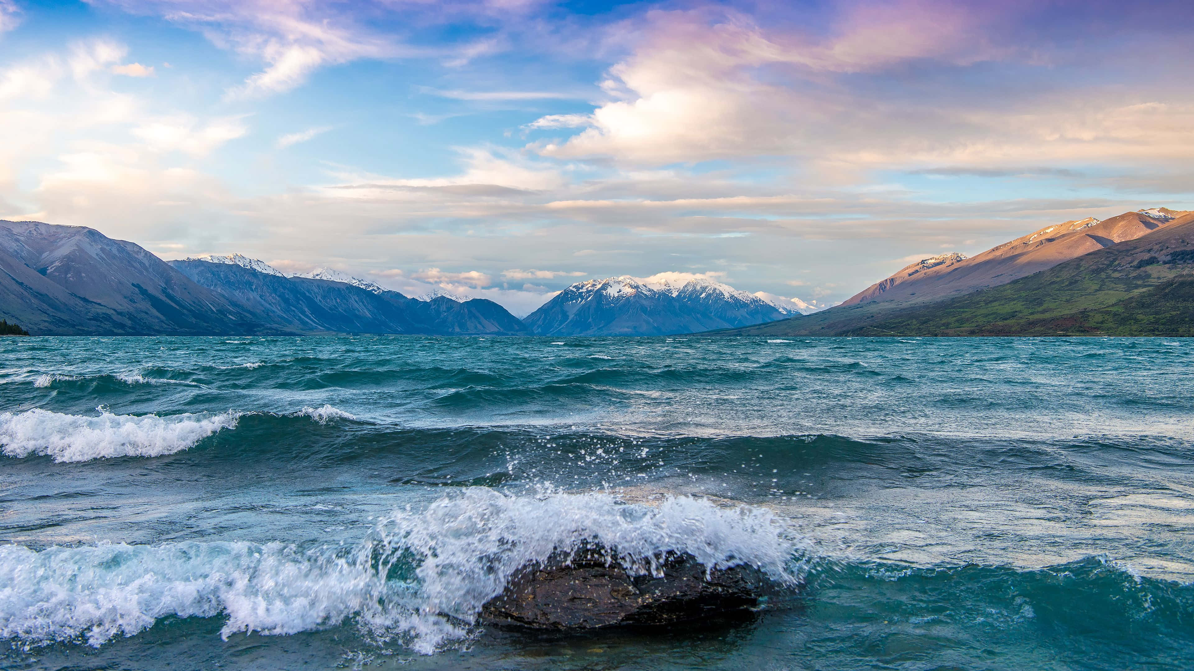 海景 桌布 壁紙 — 綠松石色的海浪拍打岩石，前方是積雪覆蓋的山脈，背景是柔和粉彩色的日落天空。