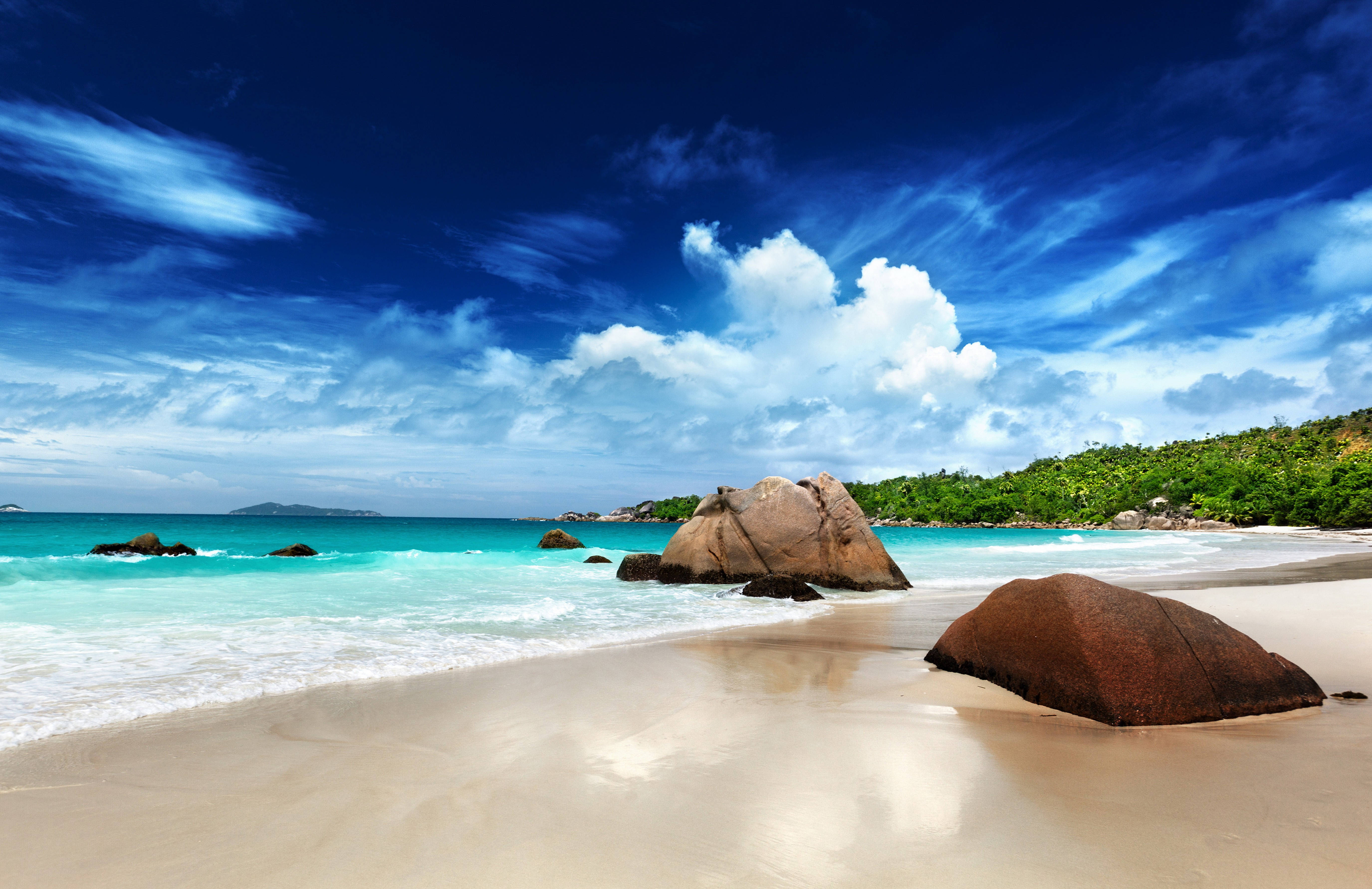 Seychelles 桌布 壁紙 — 綠松石色的海浪拍打著原始沙灘，沙灘四周環繞著巨石和鬱鬱蔥蔥的綠色山丘，上方是湛藍的天空和蓬鬆的白雲。