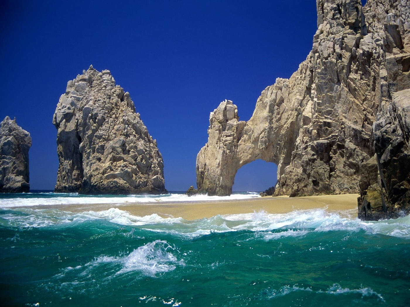 Cabo 桌布 壁紙 — 綠松石色的海浪拍打著沙灘，在廣闊的藍天下，聳立的岩石 formations 包括一個天然拱門，構成框架。