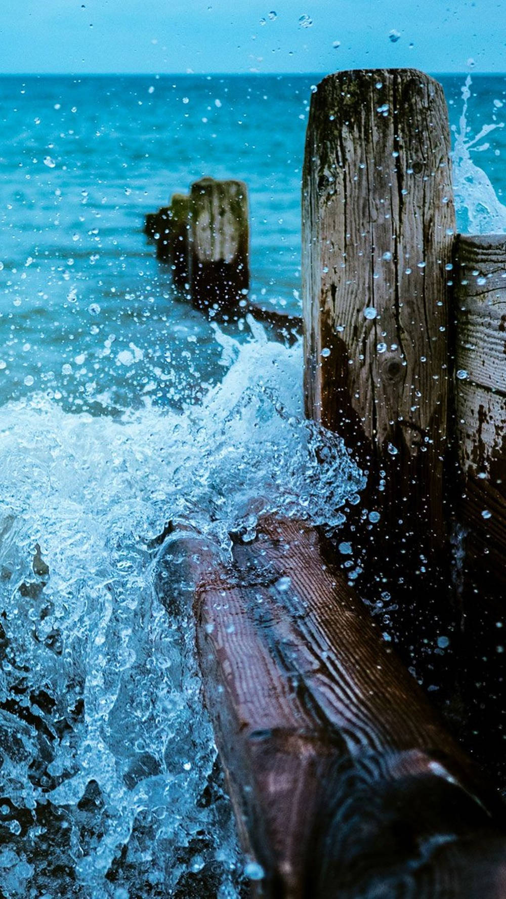 Ocean iPhone 桌布 壁紙 — 綠松石色的海浪猛烈拍打著風化的木樁，激起動態的水花。