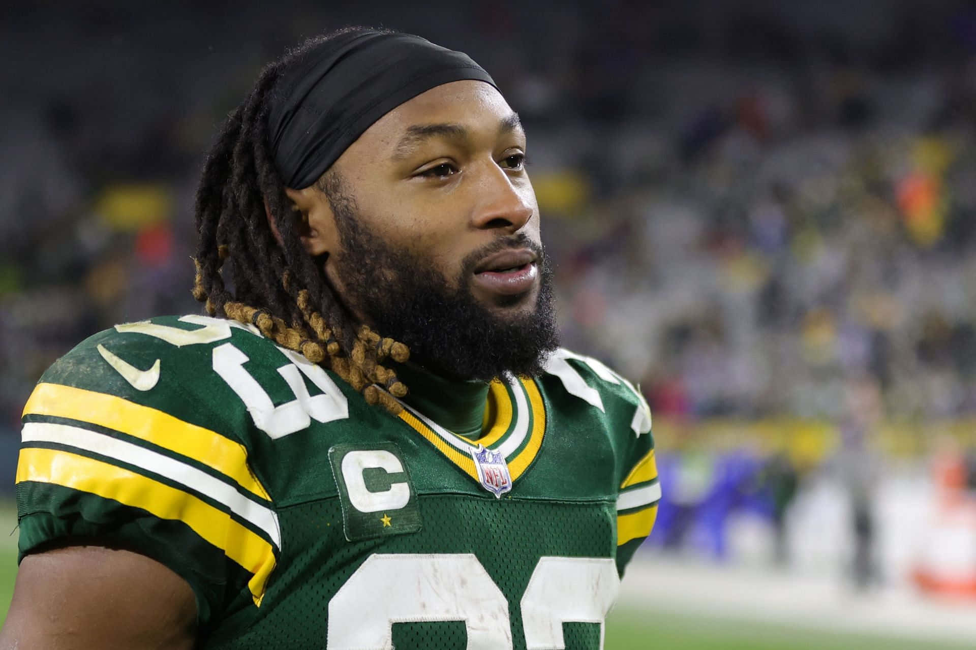 Aaron Jones 桌布 壁紙 — 綠灣包裝工美式足球員的特寫，身穿綠色和金色制服，帶有隊長 'C' 臂章，在球場上專注地凝視。