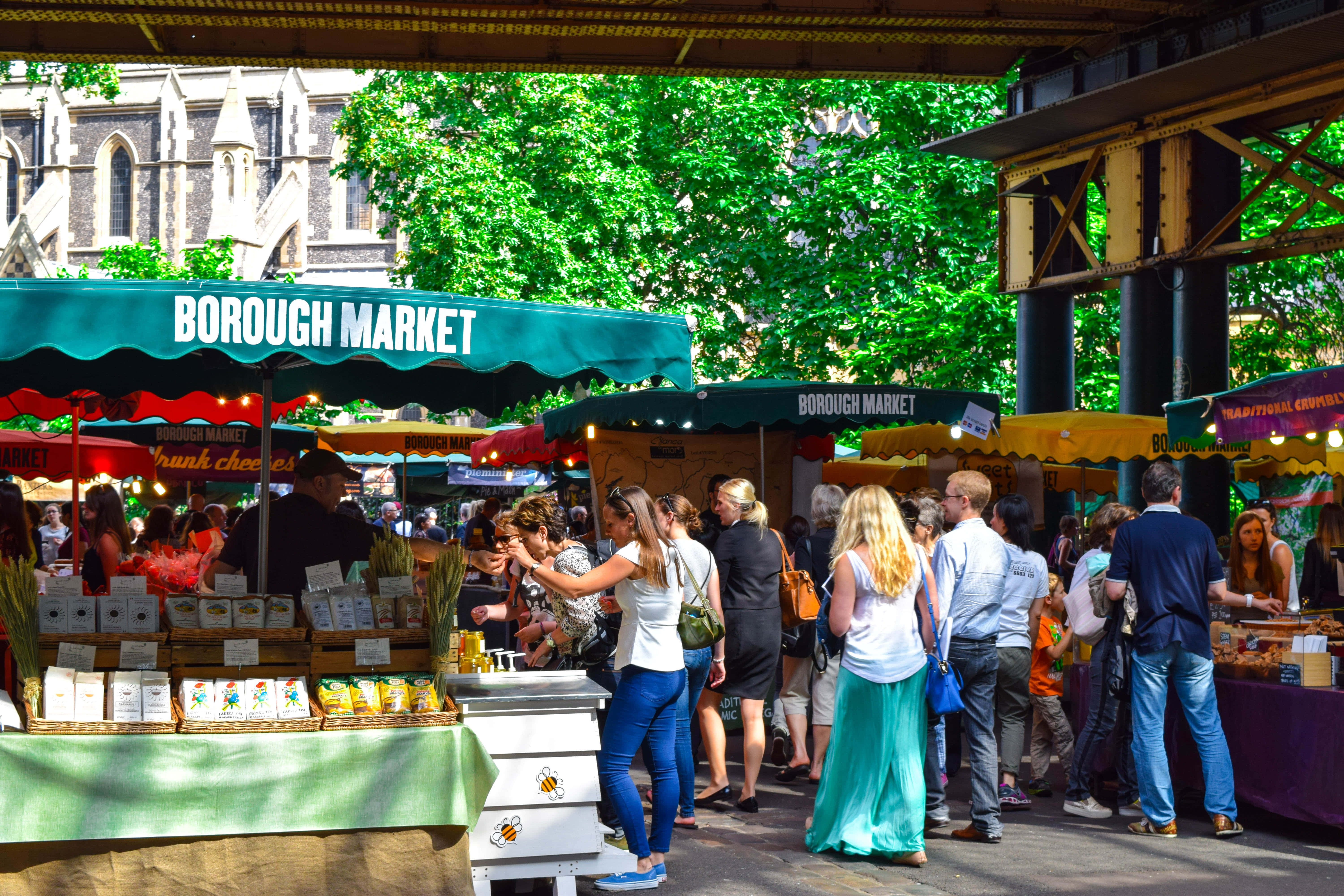Farmers Market 桌布 壁紙 — 綠色簷篷下擠滿人的波羅市場攤位，購物者在明媚夏日陽光下瀏覽食物。