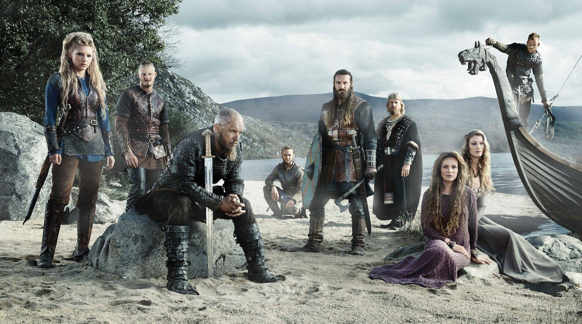 Vikings 桌布 壁紙 — 維京船員在陰天下的岩石海灘上，戲劇性地在一艘長船旁擺姿勢。