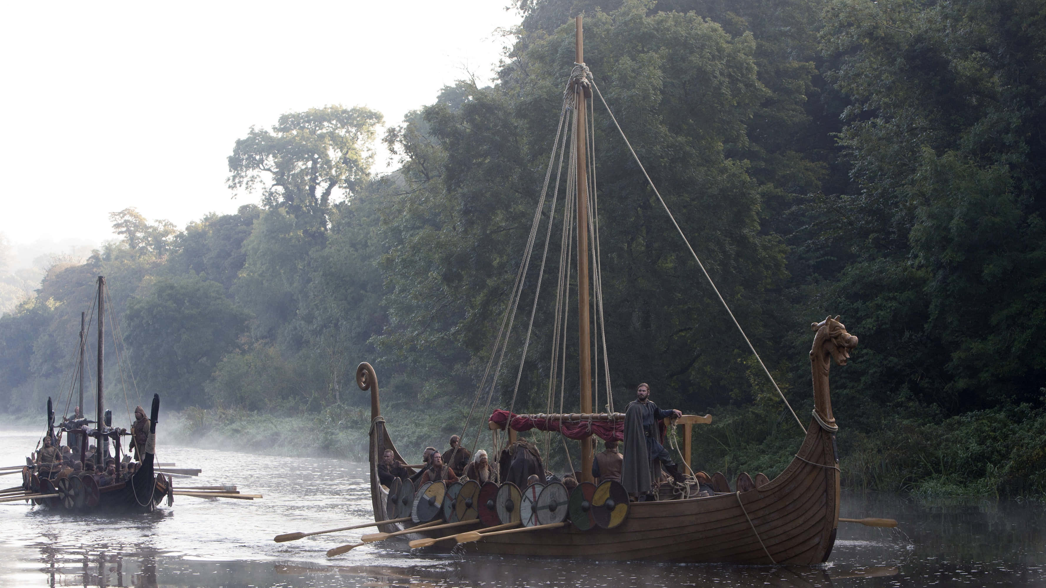 Vikings 桌布 壁紙 — 維京長船帶有龍頭，在柔和黎明光芒下，滑行於被茂密綠色森林環繞的霧氣河流中。