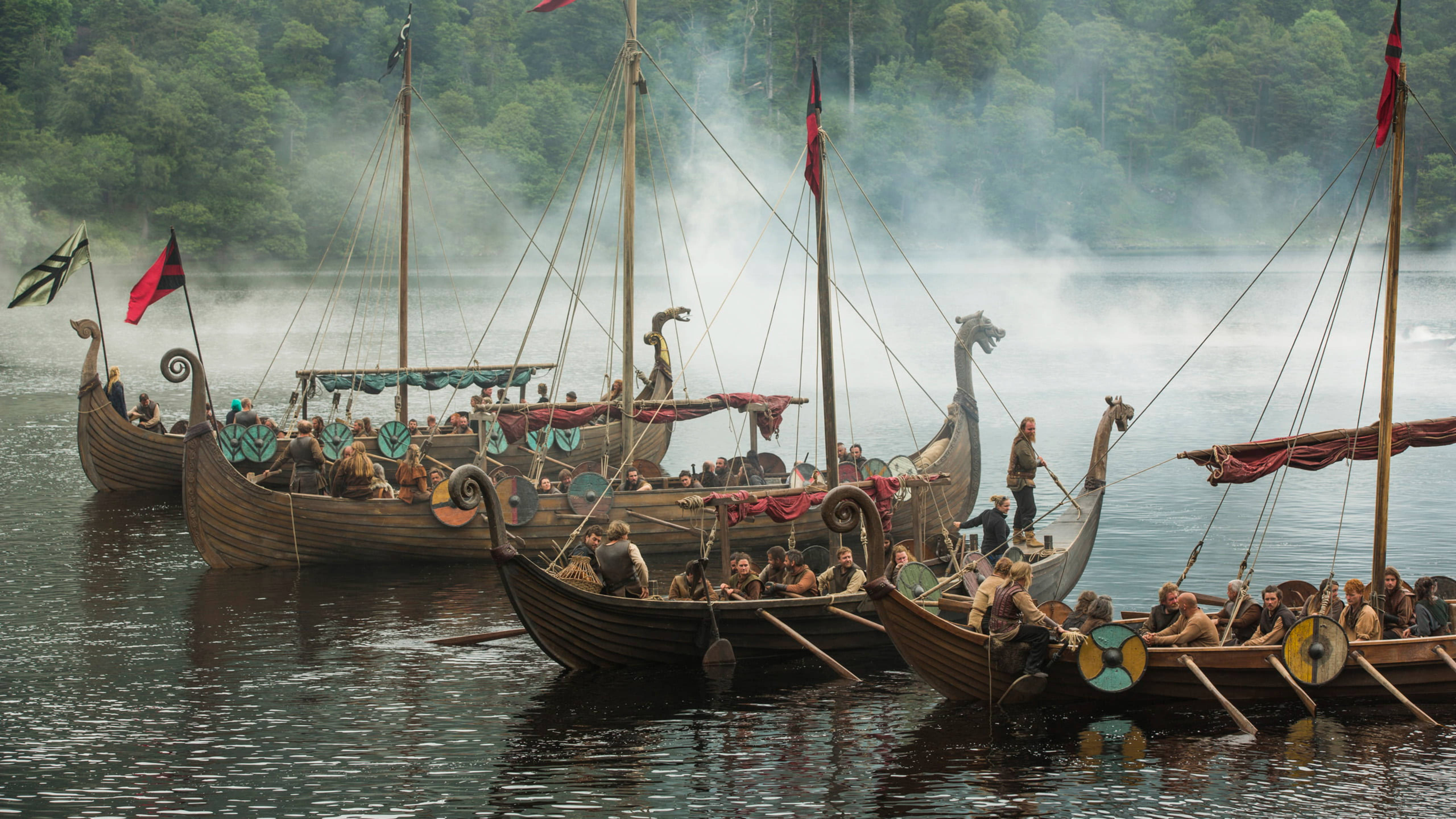 Viking Ship 桌布 壁紙 — 維京長船帶有龍頭和紅旗，在霧氣瀰漫的湖泊上行駛，周圍是茂密的綠色森林。
