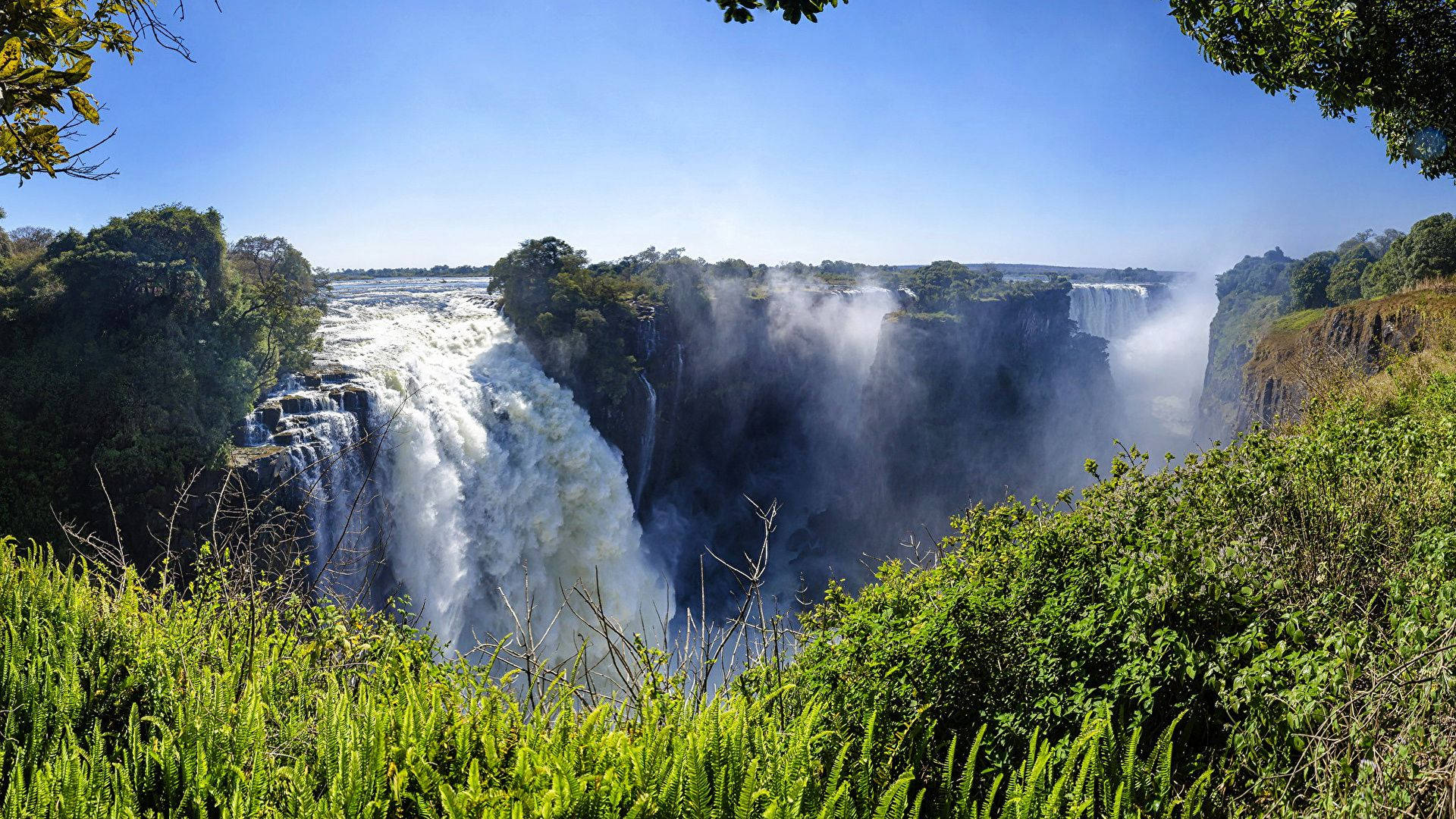 Zimbabwe 桌布 壁紙 — 維多利亞瀑布在晴朗藍天下，氣勢磅礴地傾瀉入被茂密綠葉環繞的霧氣峽谷，景色令人嘆為觀止。