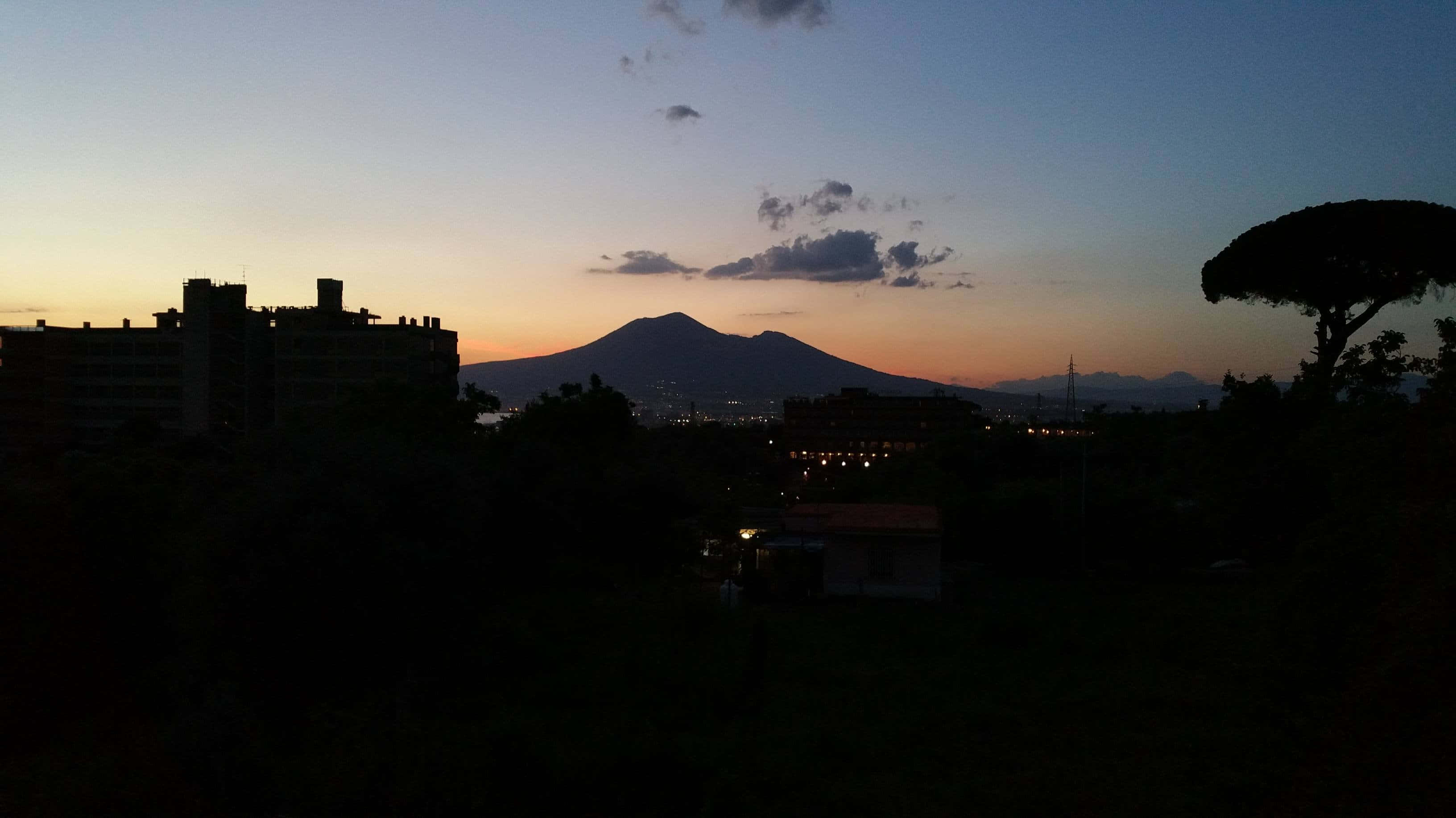 Volcano 桌布 壁紙 — 維蘇威火山的剪影在溫暖橙色地平線和烏雲的暮色城市景觀上空升起。